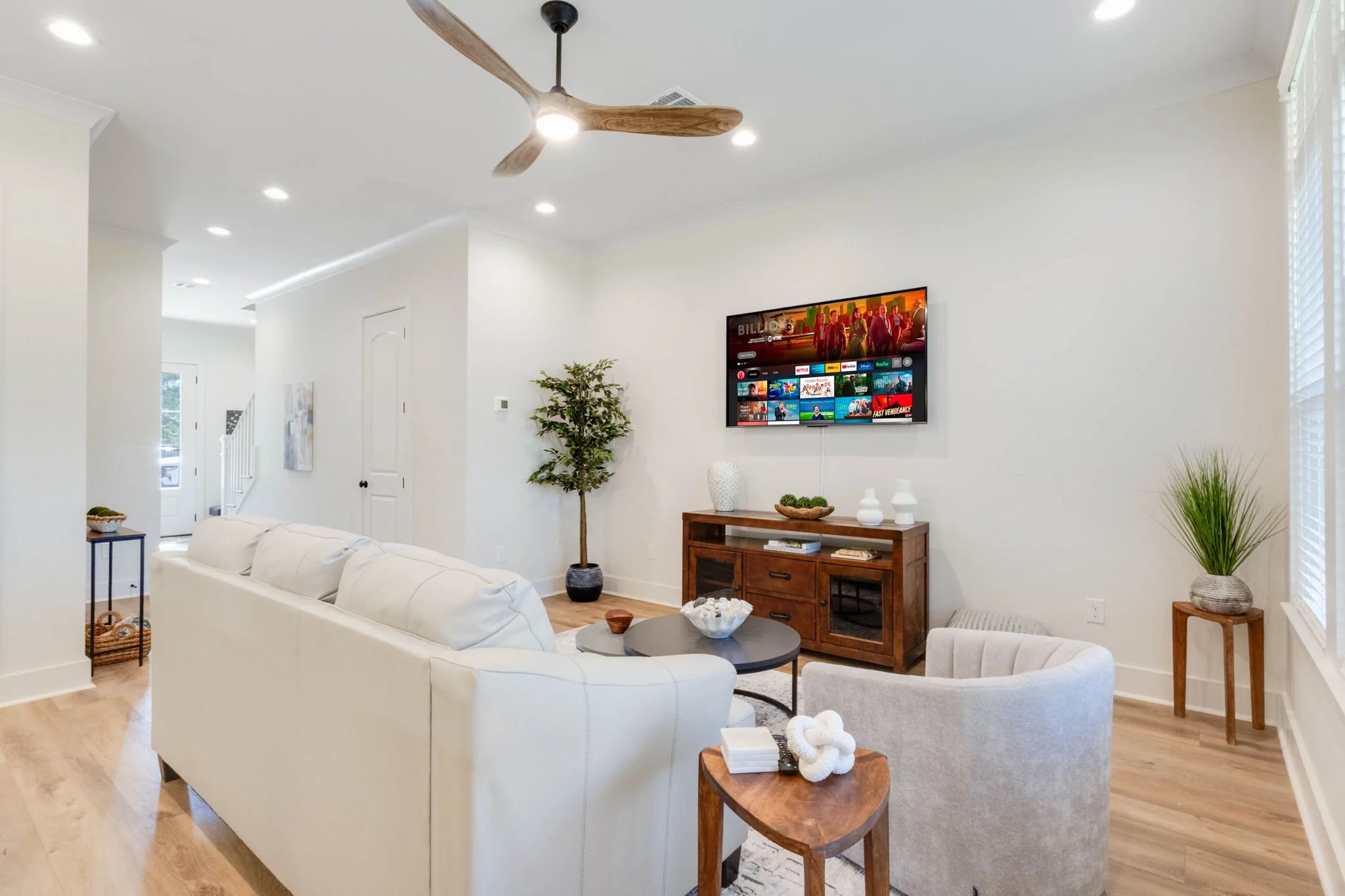 Modern living room with white sofa, gray armchair, wooden coffee table, TV mounted on the wall, and hardwood floors, decorated with potted plants and decorative vases.