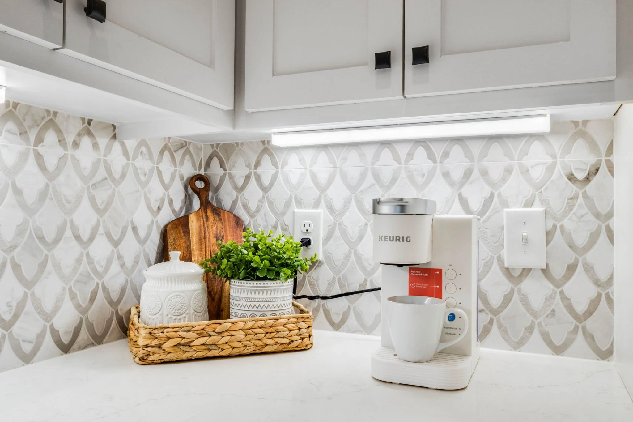 Keurig coffee maker with a white mug, a potted green plant, a wooden cutting board, and a decorative white container on a white kitchen counter with patterned tile backsplash and white cabinets above.