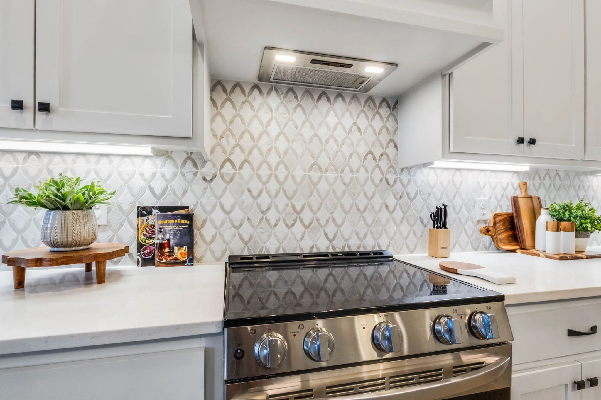 Kitchen with white cabinets, patterned backsplash, stainless steel stove, and wooden accents including cutting boards, trays, and plant pots.