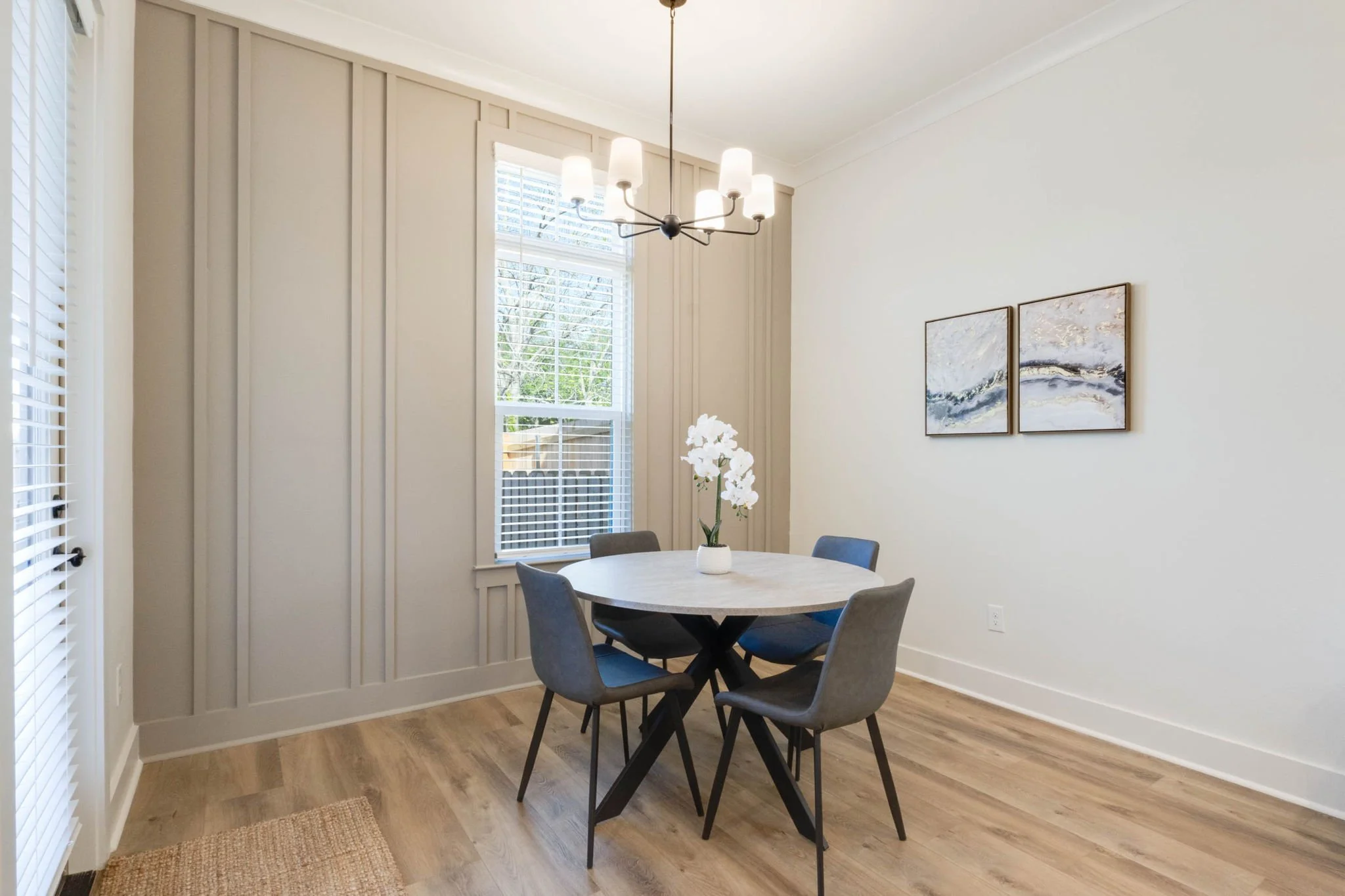 Modern dining room with a round white table and five gray chairs. A vase with white orchids is on the table. A large window with white blinds lets in natural light. A chandelier with six white lights hangs above the table. Two abstract paintings are