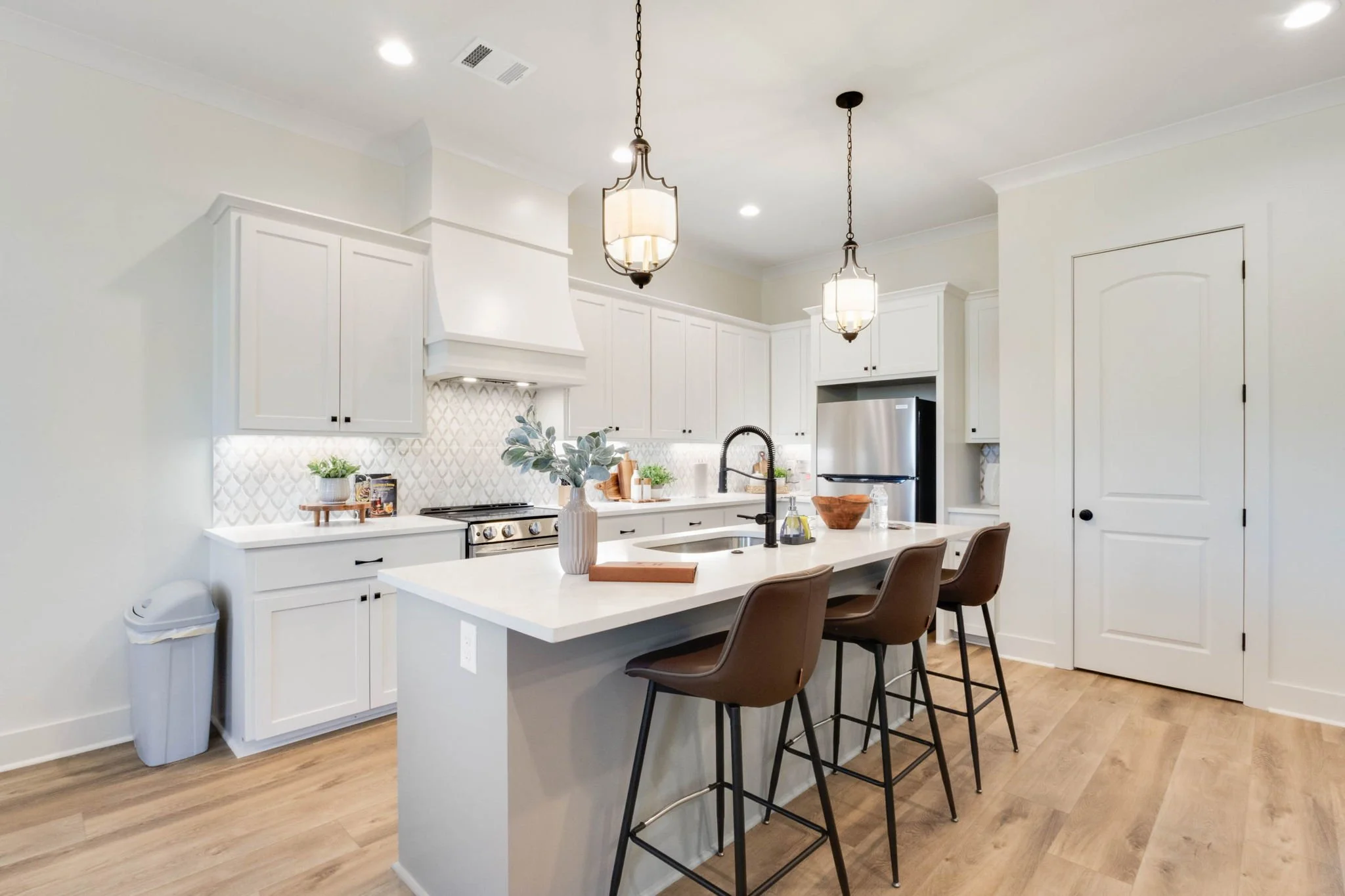 Modern white kitchen with island, brown bar stools, stainless steel refrigerator and oven, white cabinetry, pendant lighting, and wood flooring.
