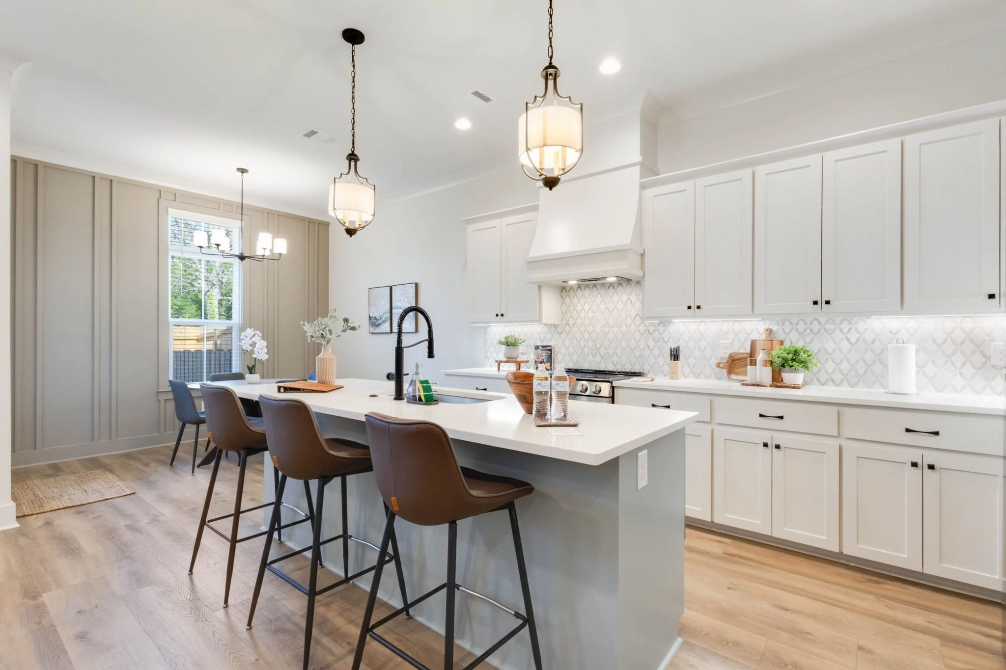 Bright kitchen with white cabinets, a large kitchen island with brown chairs, pendant and chandelier lighting, and a dining area with a window and artwork.