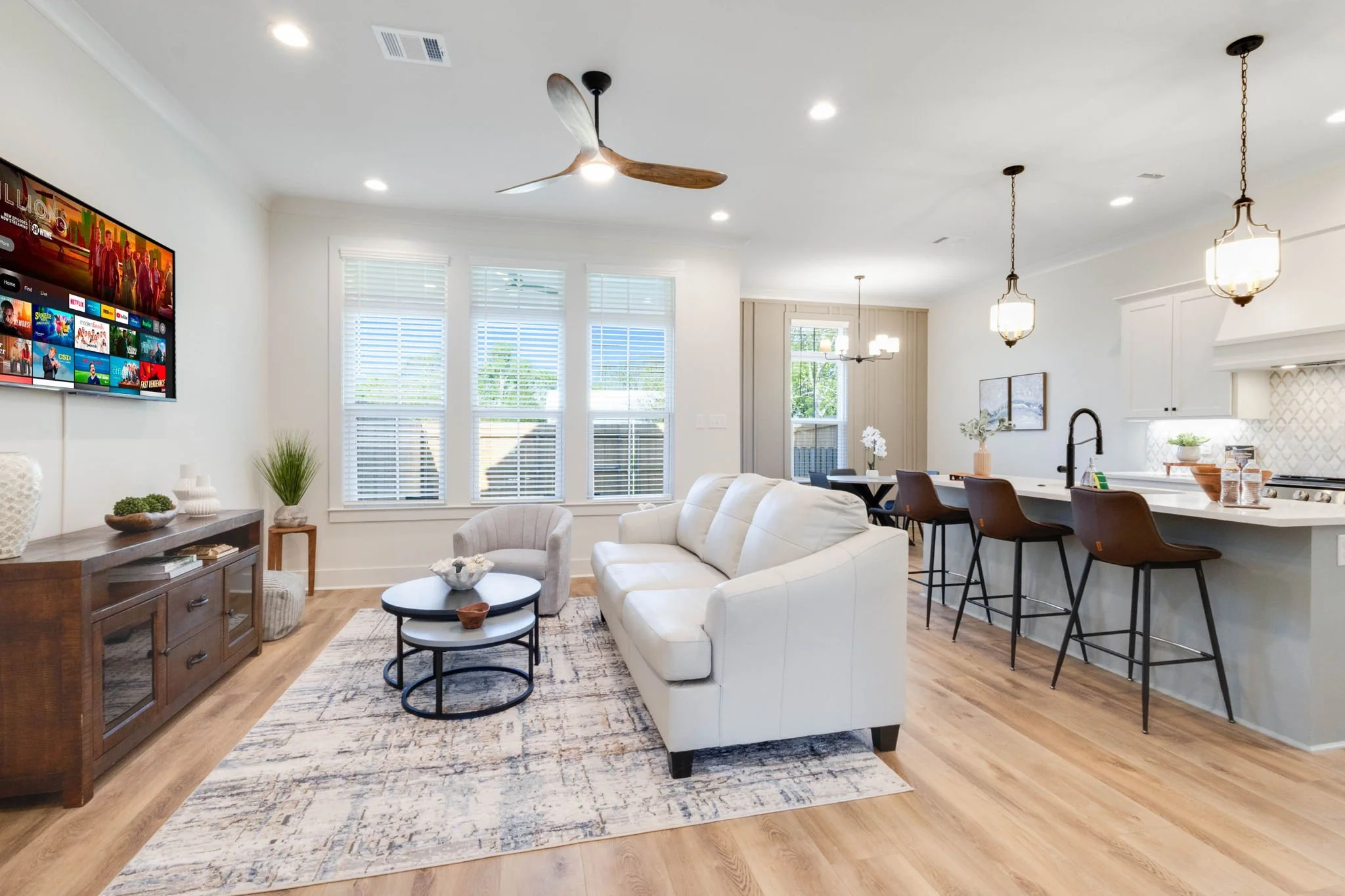 Bright, modern living room with white walls, hardwood floors, a white sofa, a small armchair, a wooden sideboard, and a wall-mounted TV. Large windows let in natural light, and open to a dining area and kitchen with pendant lighting and bar stools.