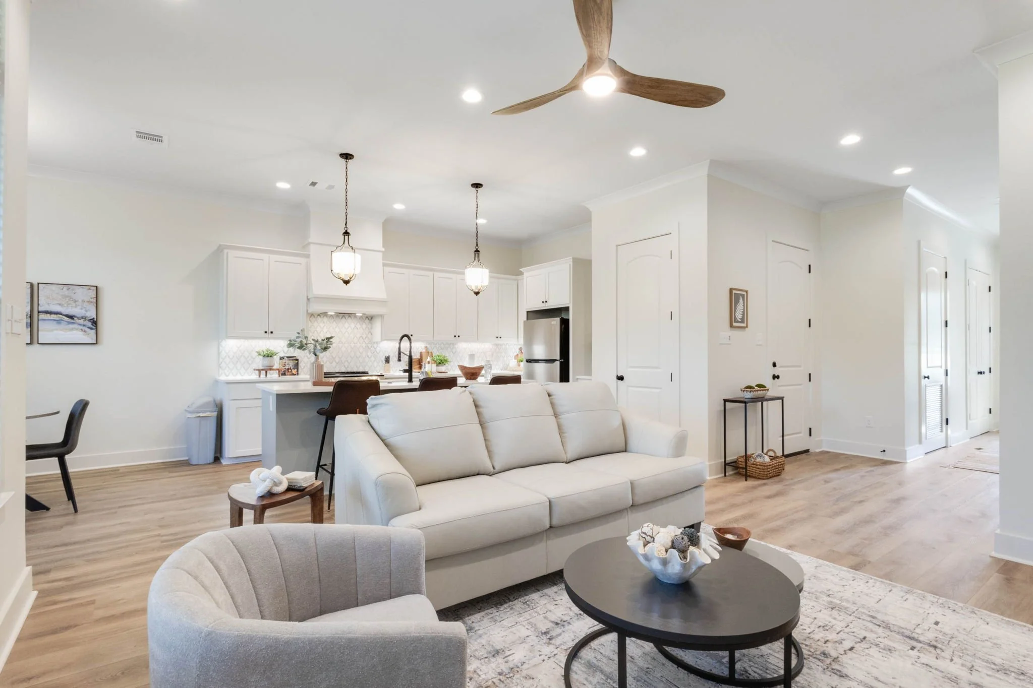 Open-concept living room and kitchen with white walls, wooden flooring, white sofas, a black coffee table with decorative items, and kitchen cabinets, a island with bar stools, pendant lighting, ceiling fan, and various decorative elements.