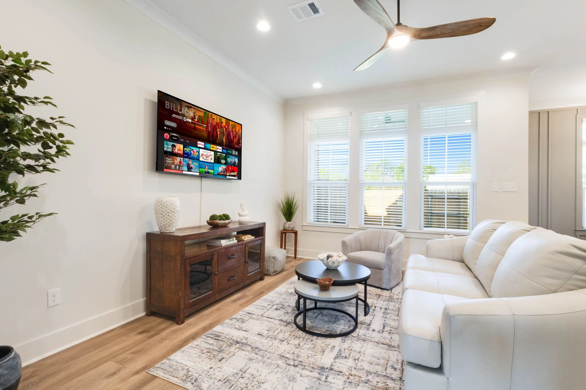Bright living room with white walls, hardwood floors, a white sofa, a beige armchair, a black round coffee table, a wooden TV stand, a flat-screen TV, and large windows with white blinds and views of trees outside.