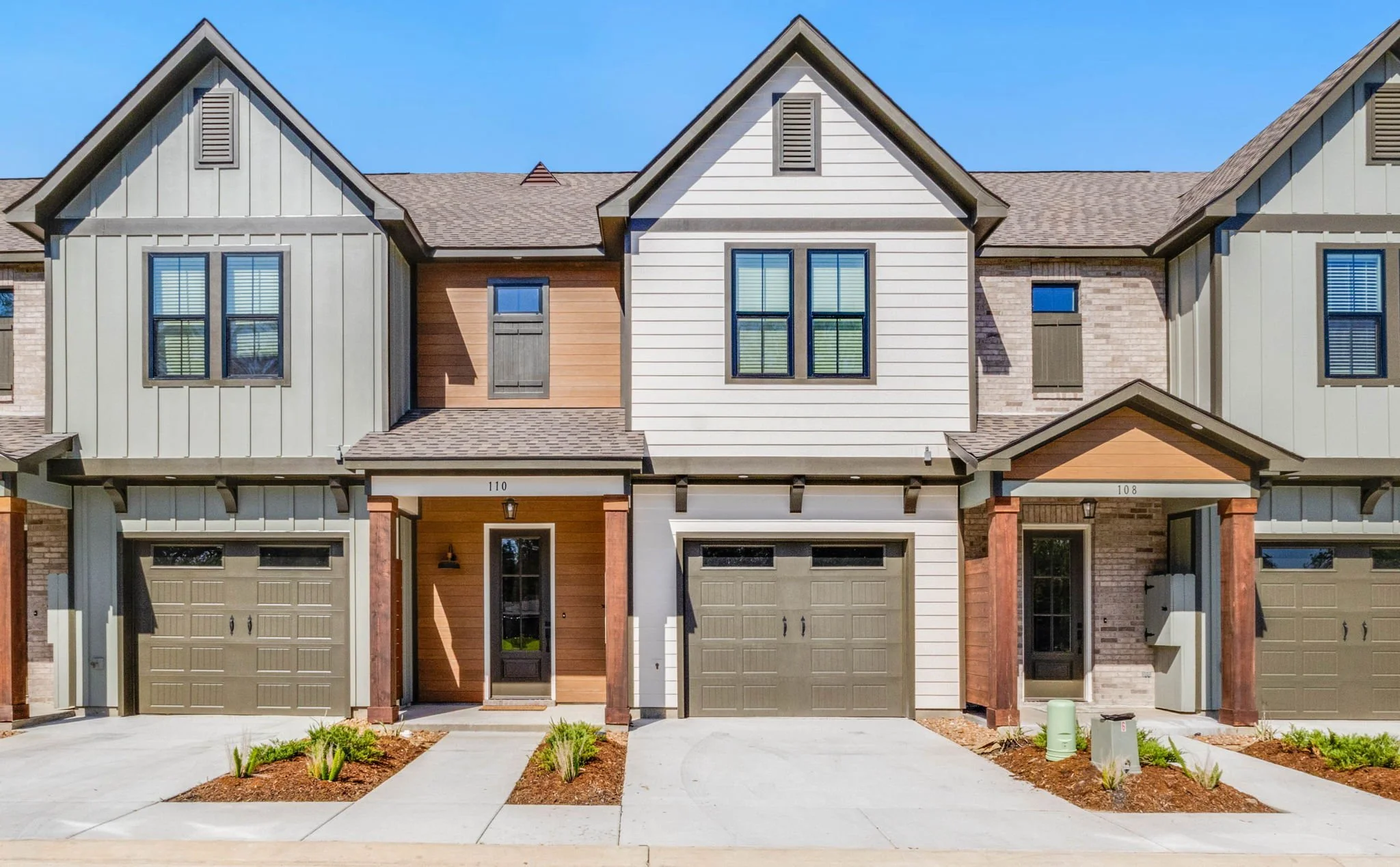 Modern townhouses with attached garages, painted in neutral colors, with small front yards and landscaping, under a clear blue sky.