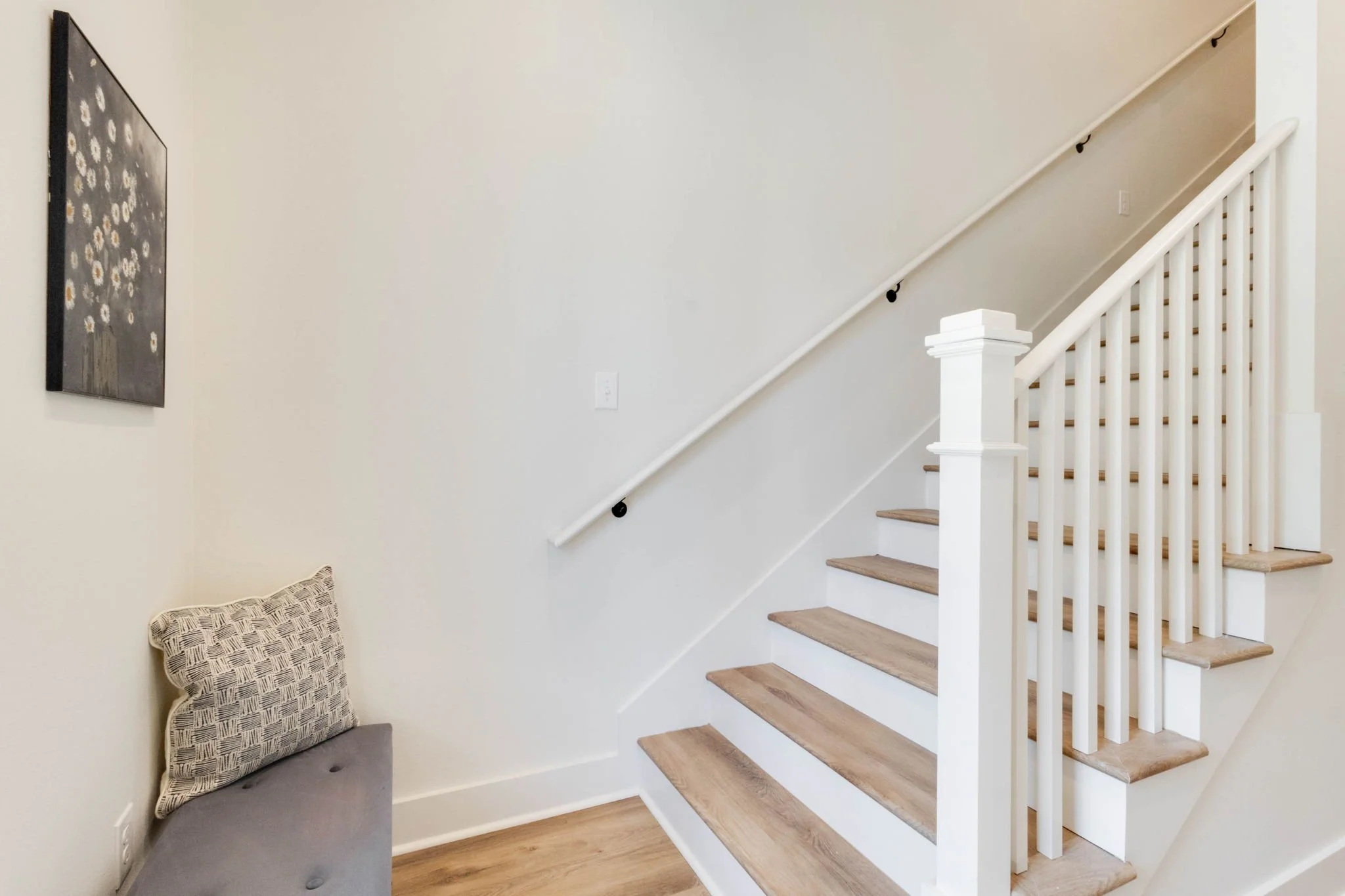 Interior view of a staircase and a small seating area with a cushion, white walls, and a wall art depicting white flowers.