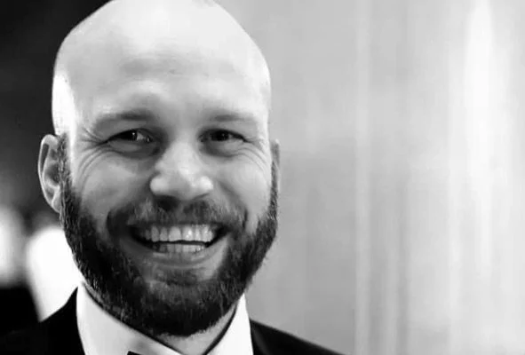 A smiling man with a beard and bald head in a formal setting, wearing a suit and tie.