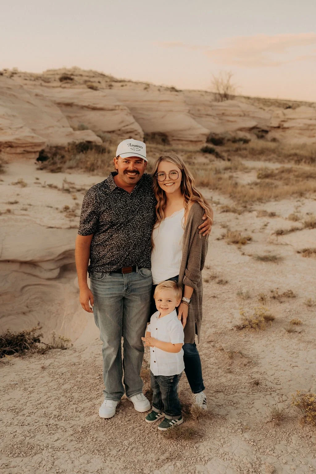 A family of three enjoying time together outdoors on a desert landscape with natural rock formations and sparse vegetation. The parents and their young son stand close, smiling, under a warm late afternoon sky, capturing a candid family moment in a s