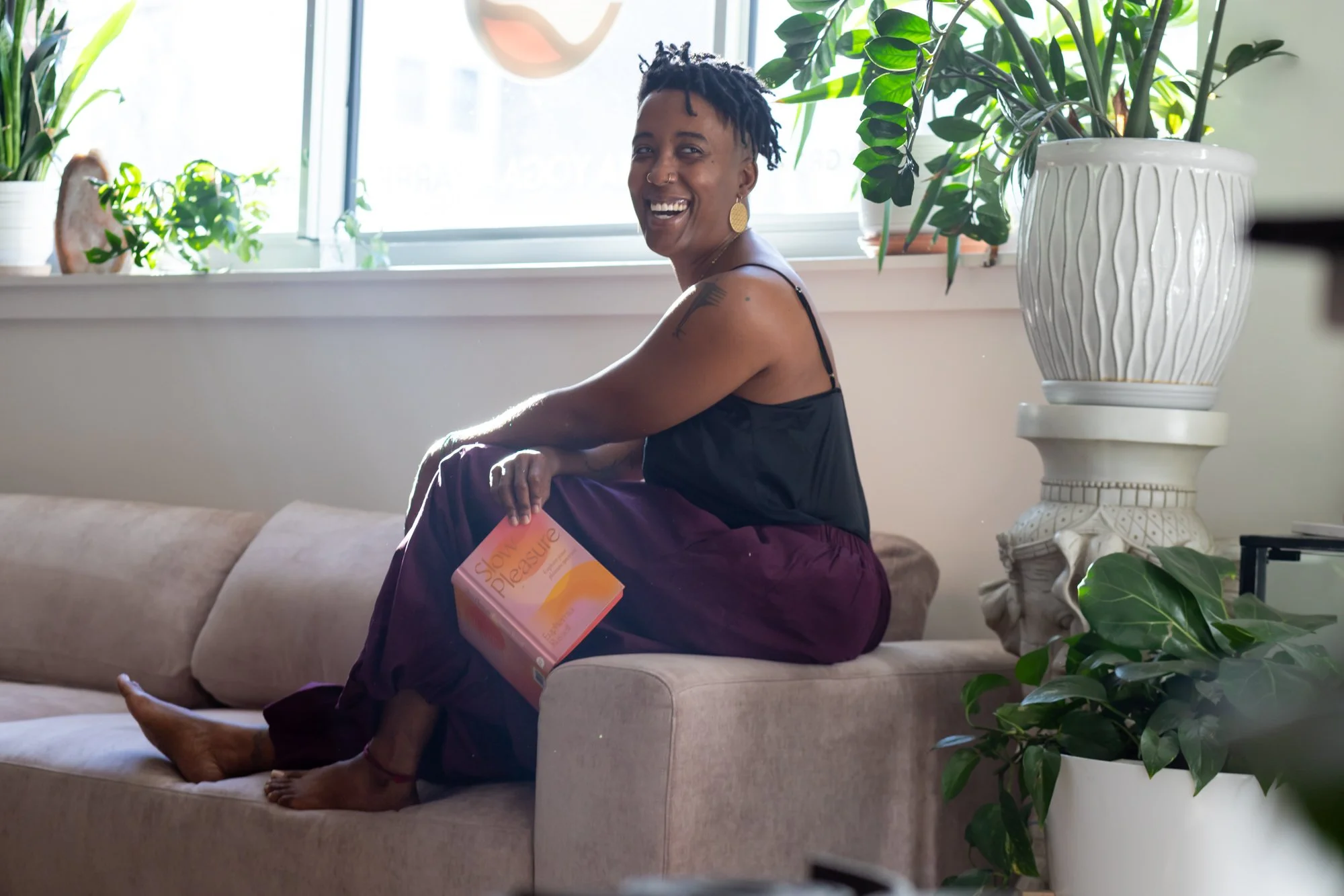 A person sitting on a beige sofa, smiling, holding a book labeled 'Slow Pleasure,' in a well-lit room with several green plants near a window.
