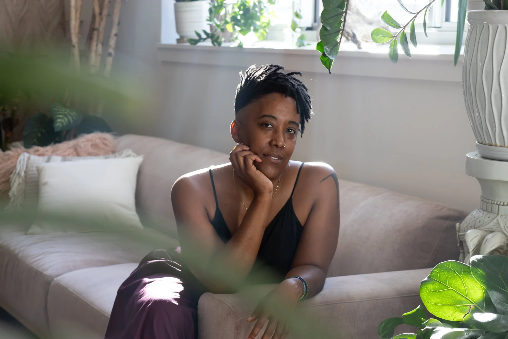 A person with short dreadlocks and a nose ring sitting on a beige couch in a sunlit room, resting their chin on their hand, with green plants and white walls in the background.