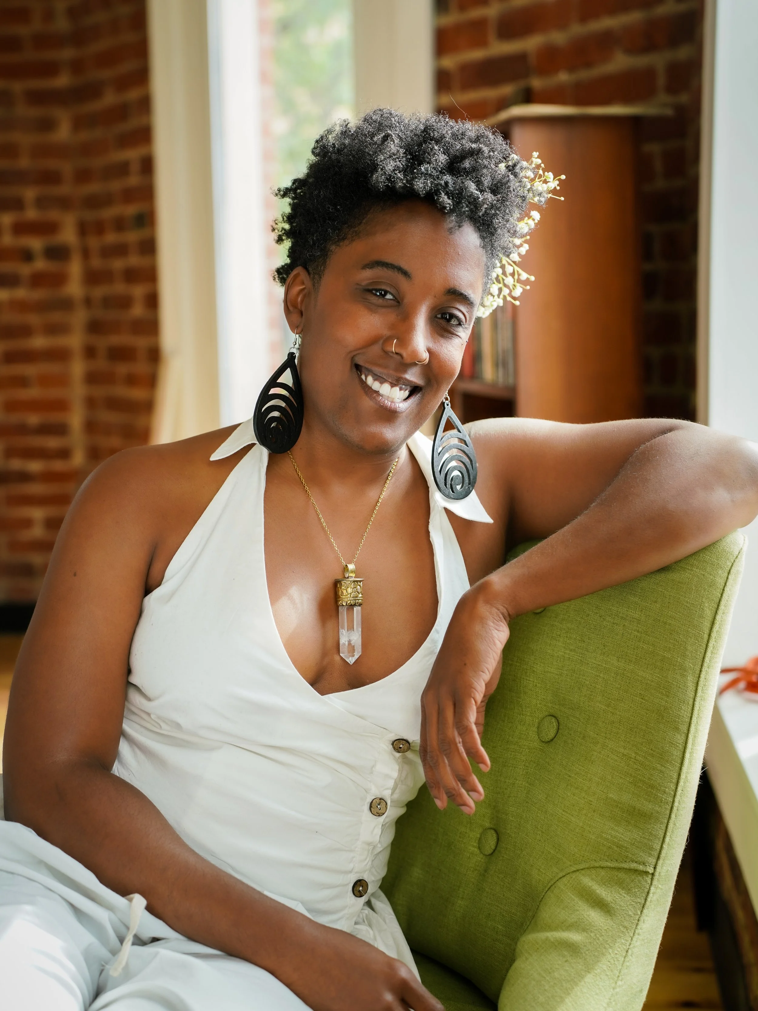 A smiling person with short curly hair, wearing large black earrings, a white clothes with buttons, and a necklace with a crystal quartz pendant, sitting on a green chair in a room with brick walls and a window.