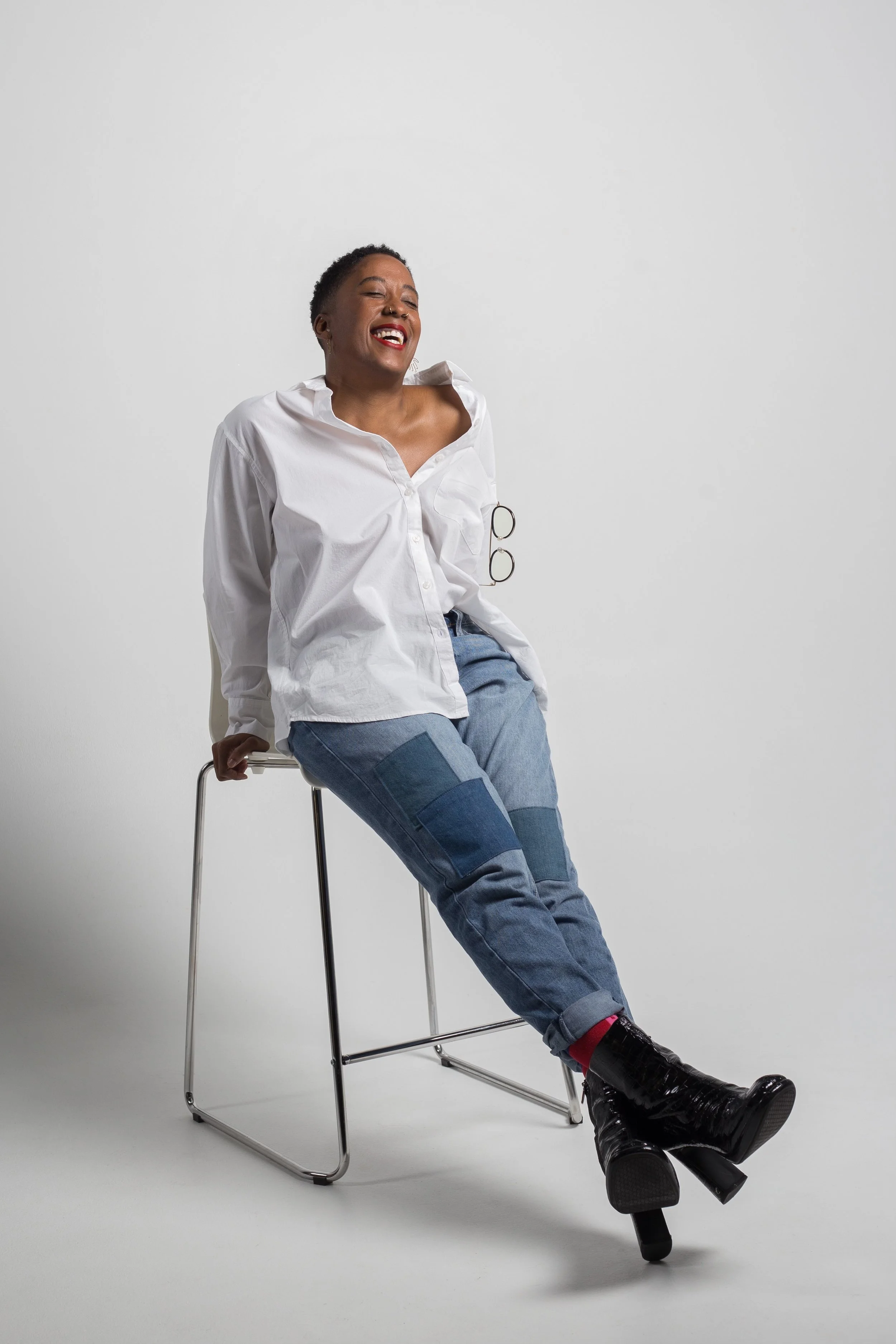 A woman sitting on a chair with a wide smile, wearing a white shirt, patchwork jeans, and black heeled boots, against a plain white background.