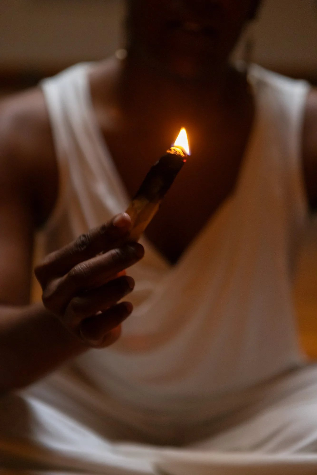 A person holding a lit palo santo in front of their chest, dressed in a white sleeveless top, in a dimly lit environment.