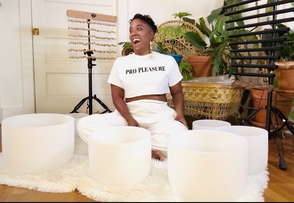 A person sitting on a white fluffy rug, surrounded by large white crystal singing bowls, in a room with houseplants, a door, and sound healing equipment in the background.
