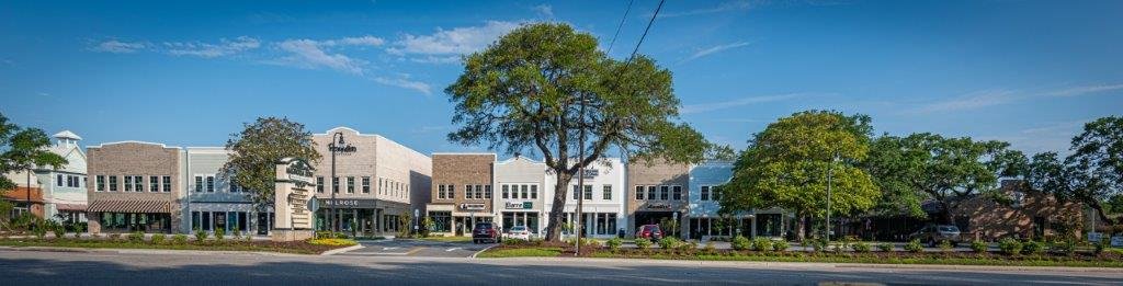 A row of small commercial buildings with storefronts on a city street, with trees along the sidewalk and a clear blue sky above.