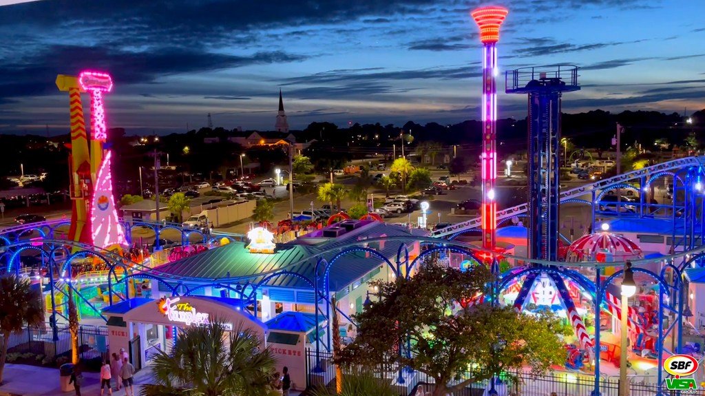 Brightly lit amusement park rides and structures at dusk with colorful neon and LED lights.