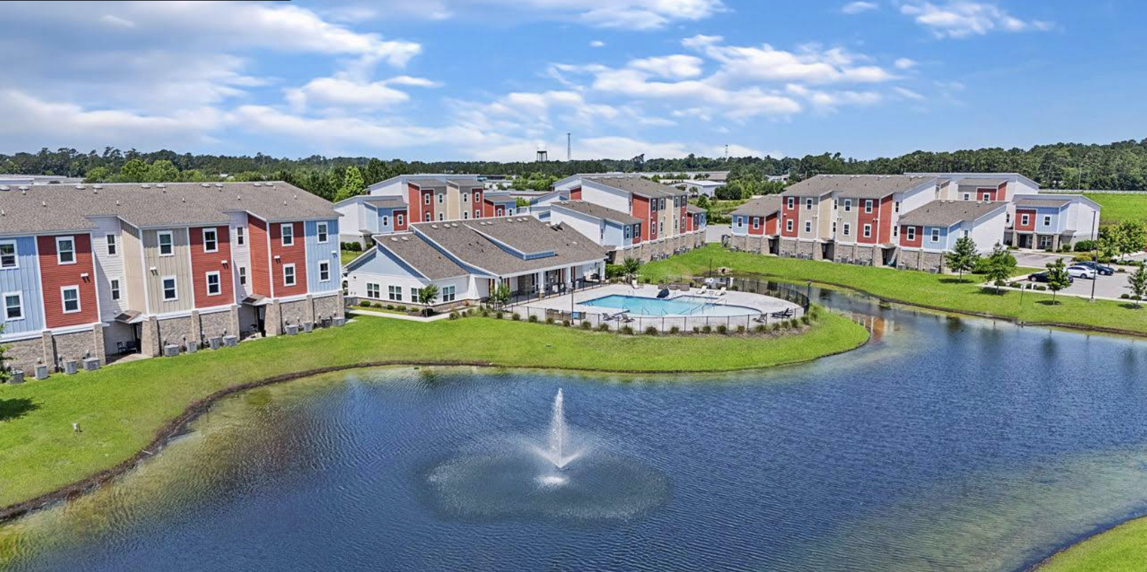 A residential apartment complex with colorful buildings surrounding a pond with a fountain, a swimming pool, and maintained green lawns under a partly cloudy sky.