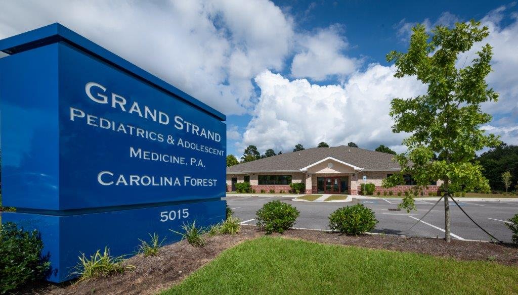 Blue sign next to a building reads 'Grand Strand Pediatrics & Adolescent Medicine, P.A., Carolina Forest, 5015'; parking lot, trees, and a partly cloudy sky in the background.