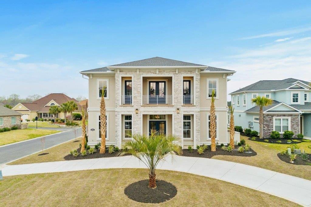 A large, two-story house with a light-colored exterior, small balcony, and surrounding tropical landscaping, situated in a suburban neighborhood.