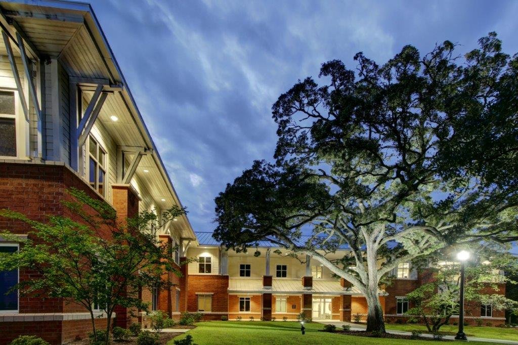 A two-story brick residential building with white trim, large windows, and a front porch, featuring a large sprawling tree with dense green foliage in front and a well-maintained lawn, under a evening sky with clouds.