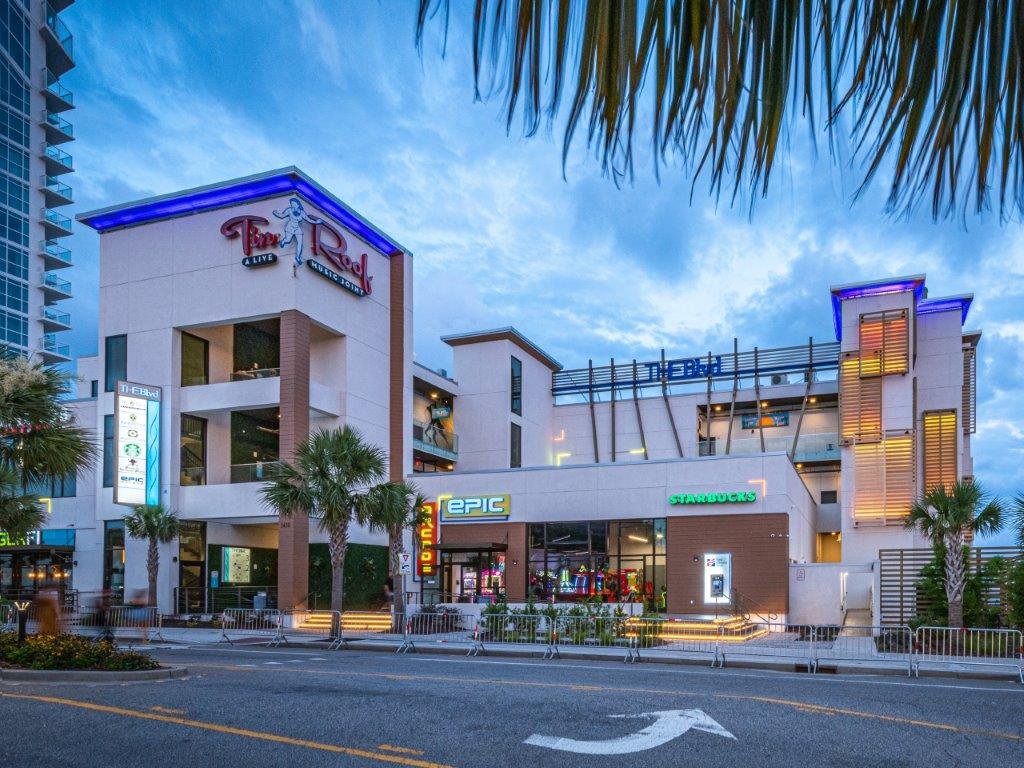 Modern multi-story shopping center with signage for Top Red, Epic, Starbucks, and T-Mobile, surrounded by palm trees, with a cloudy sky in the background.