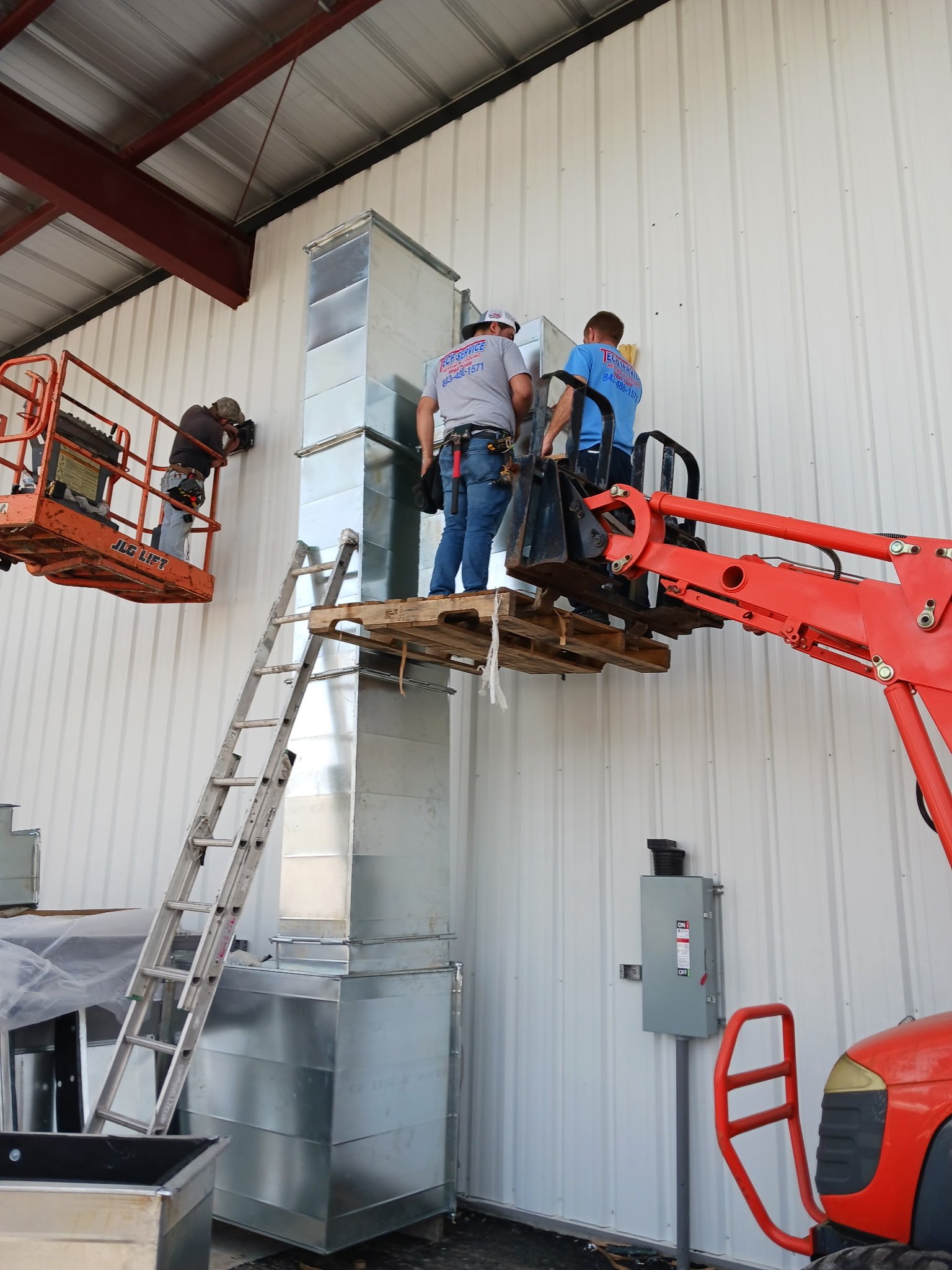 Three workers are installing or repairing ductwork on a high wall in an industrial building. Two are on a wooden platform attached to a cherry picker lift, and one is on a ladder. The wall is white metal siding, and there are various tools and equipm