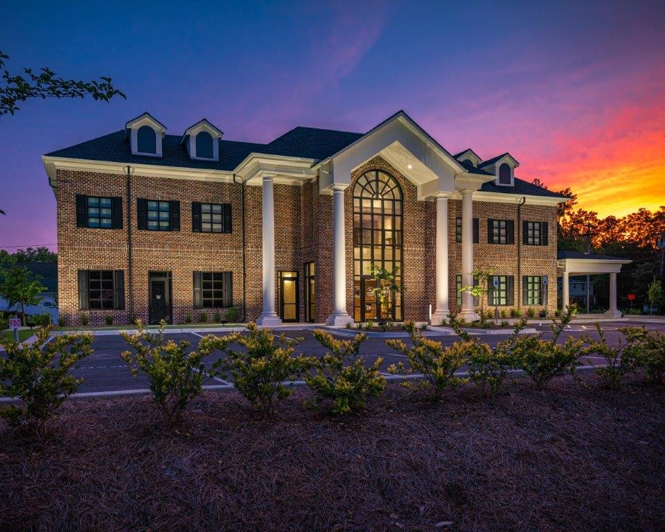 A large brick building with white columns and a tall arched window, illuminated during sunset with a colorful sky in the background.