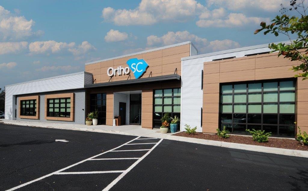 Exterior view of a modern building with a sign that reads 'OrthoSC', likely an orthopedic clinic, with a parking lot in front and blue sky with clouds above.
