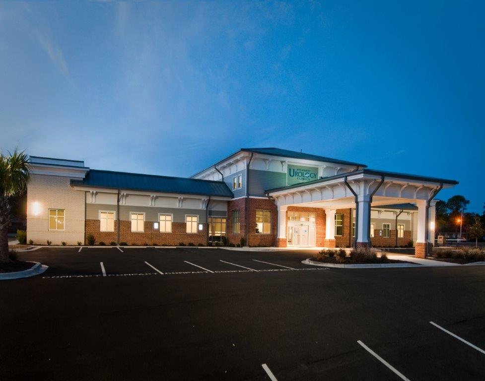 Exterior view of a modern clinic building at dusk with illuminated entrance, parking lot, and a sign displaying 'Urology Clinics'.