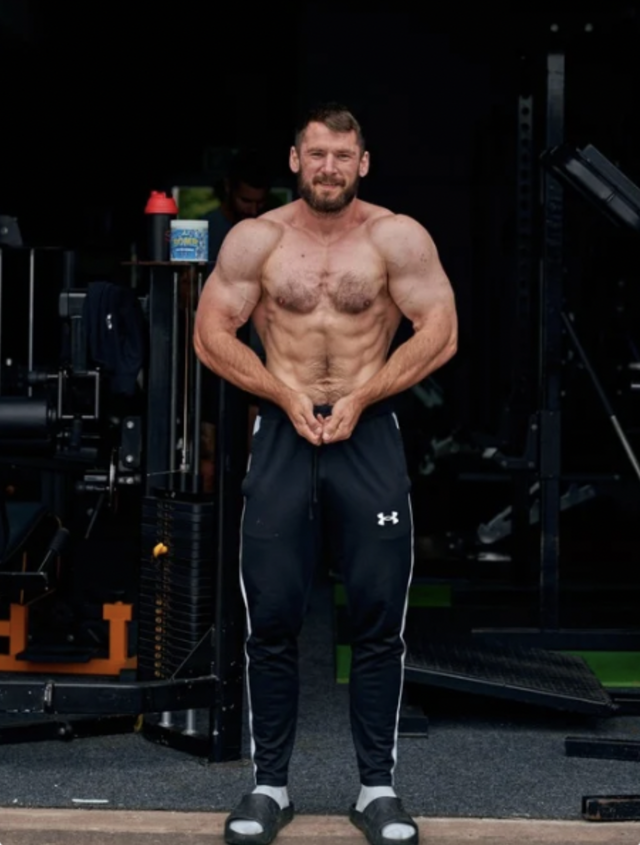 A muscular man with a beard posing shirtless in a gym, flexing his muscles and standing on a green mat.