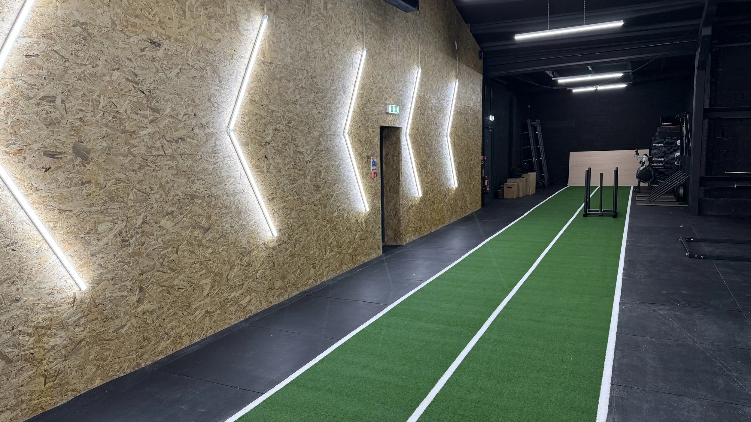 Indoor gym with black flooring, wooden wall with geometric light fixtures, green artificial turf running track with white lines, and workout equipment in the background.