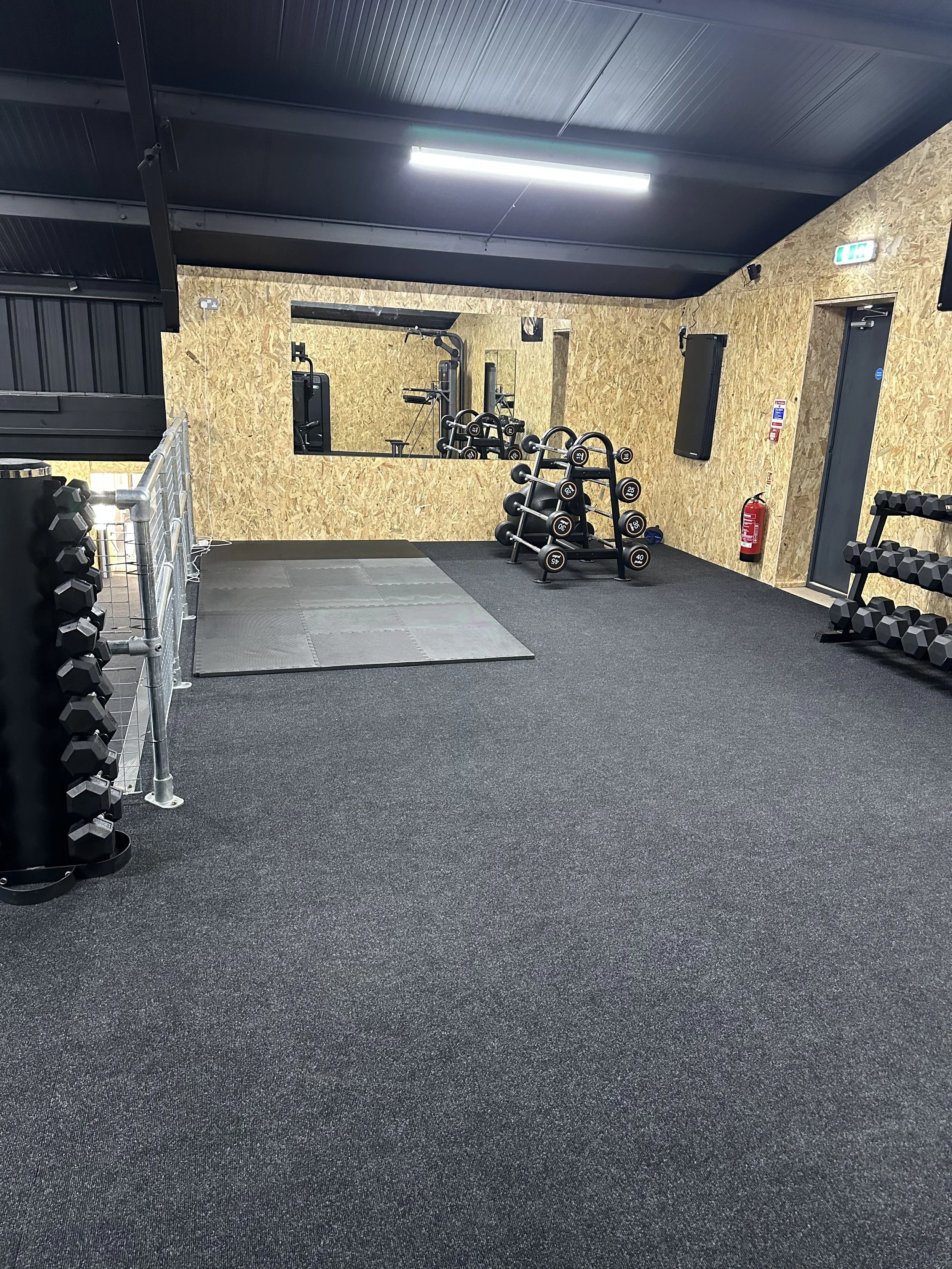 An empty gym with black rubber flooring, a mirrored wall, and various free weights and dumbbells on racks.