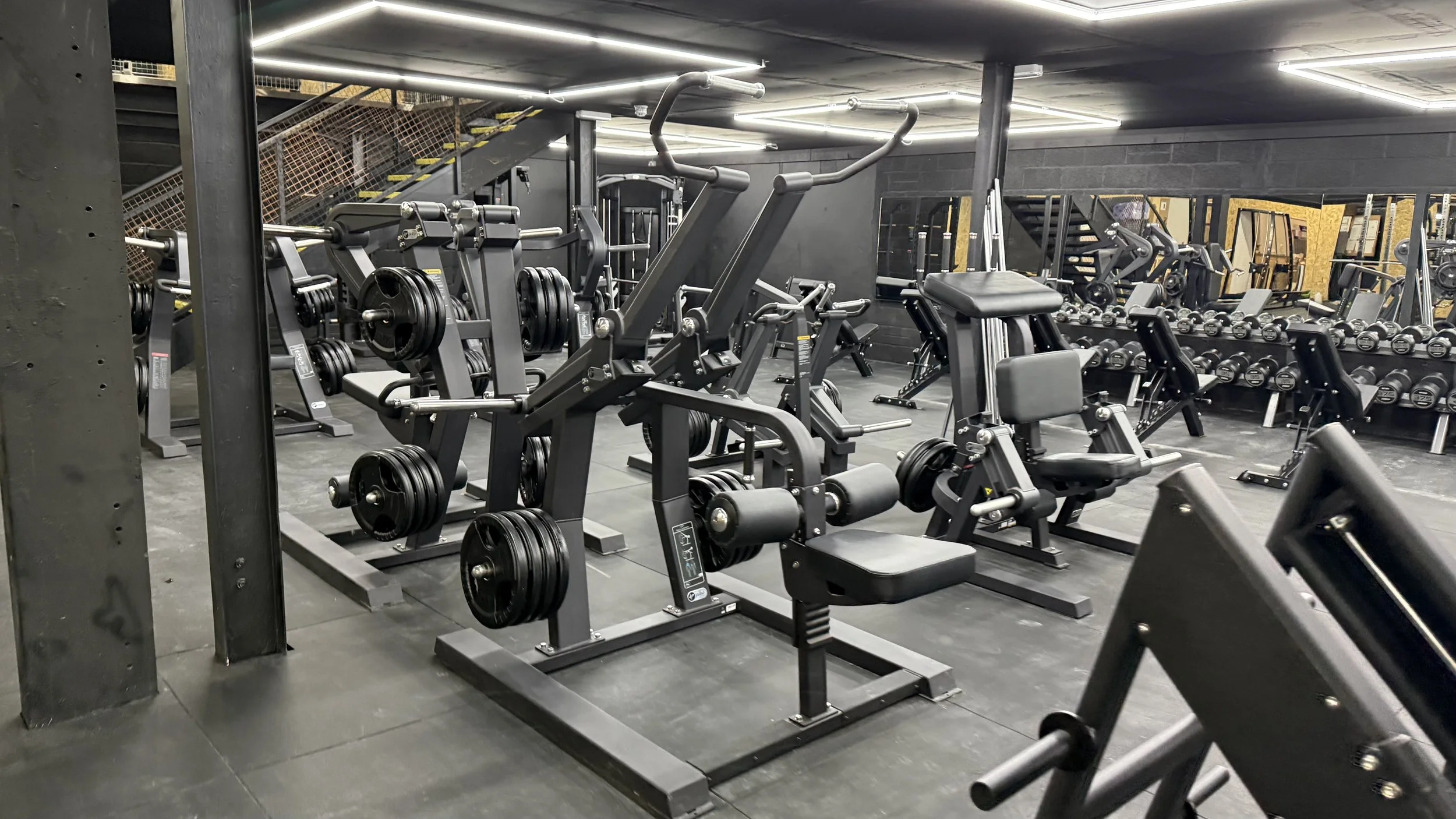 Gym equipment including weight machines, free weights, and benches in a modern gym with black walls and fluorescent lighting.