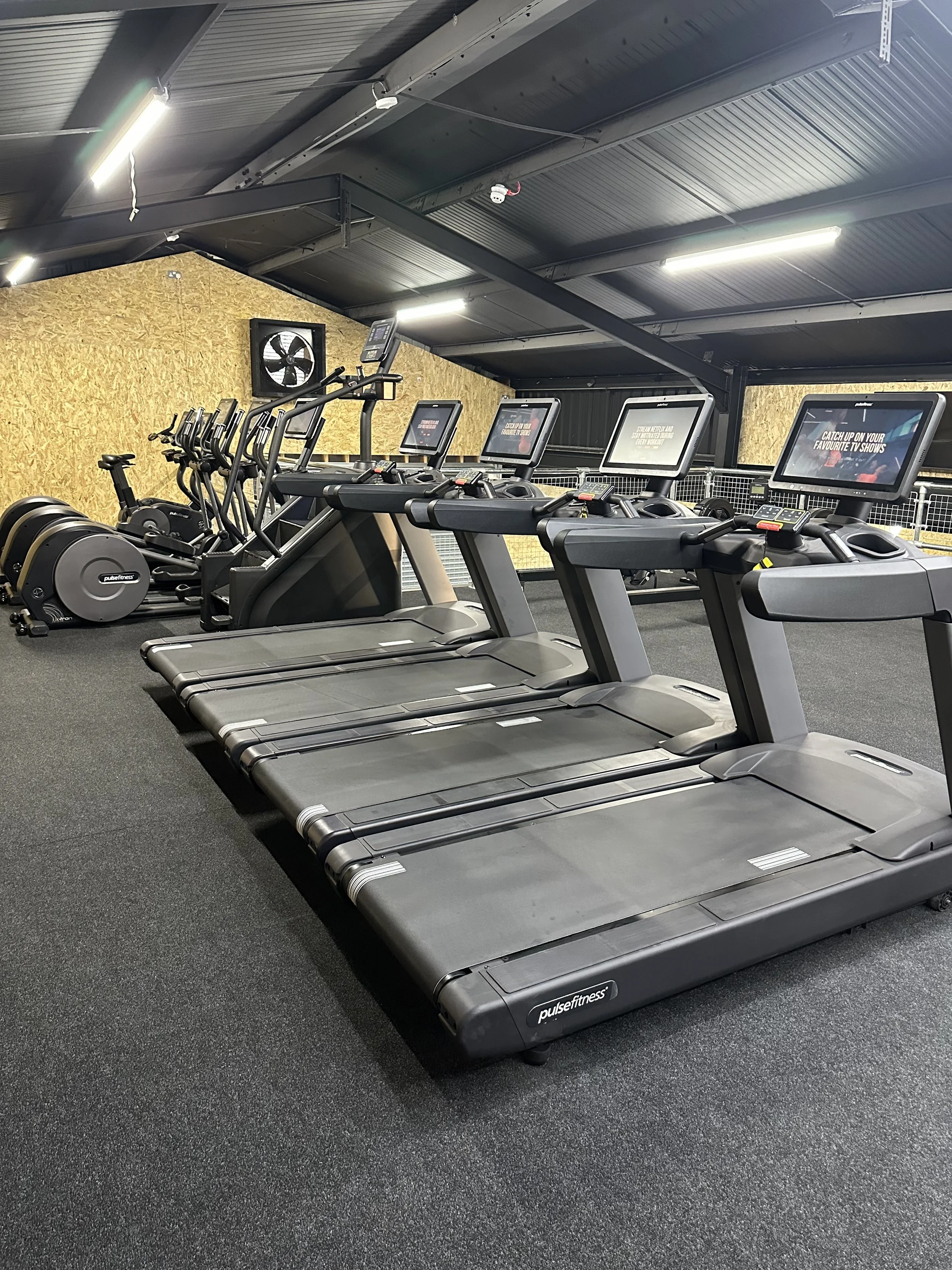 Row of treadmills and cardio equipment in a gym with black rubber flooring and yellow wooden wall in the background.