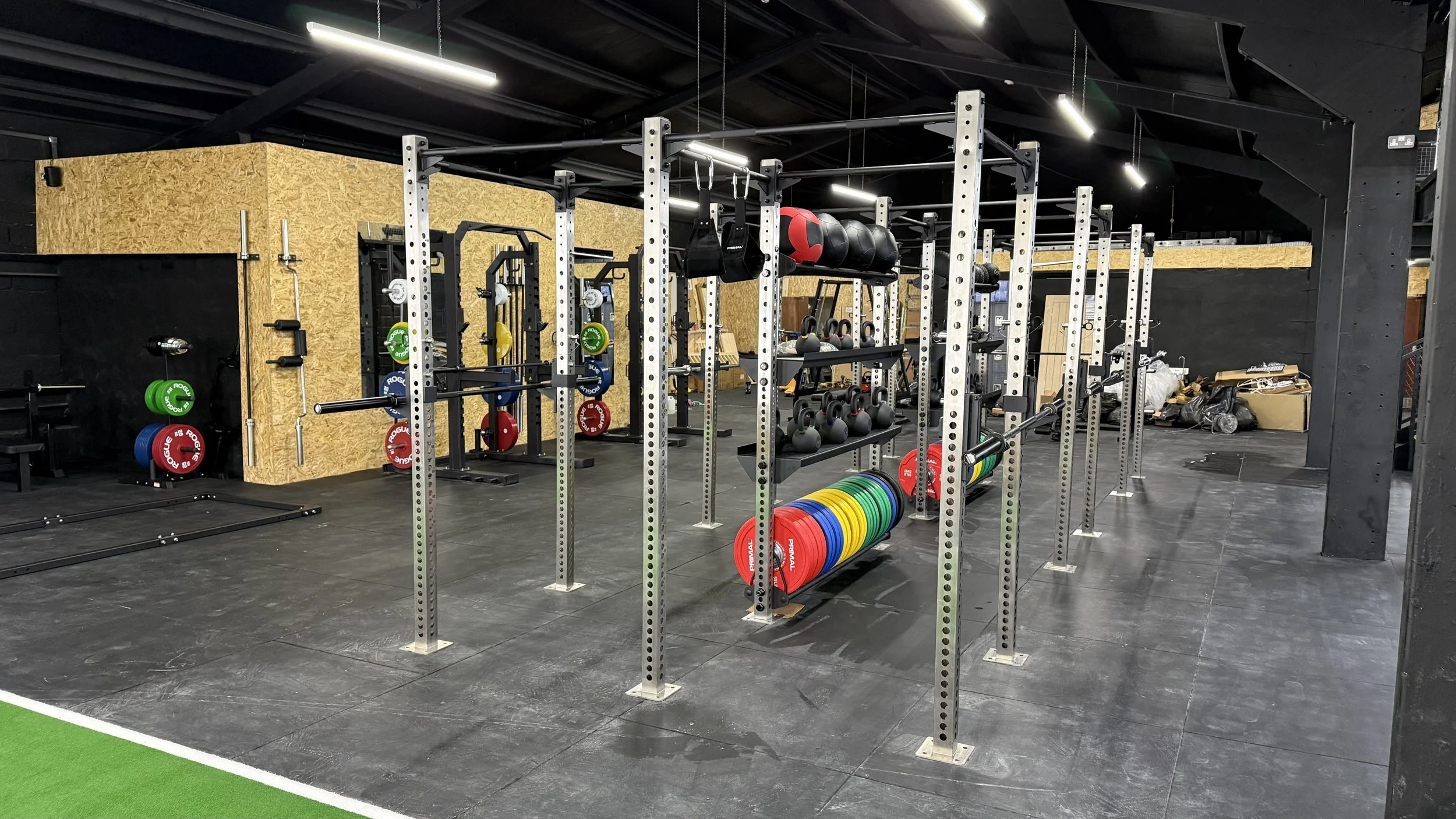 Gym with weightlifting equipment, including barbells, weight plates, kettlebells, and medicine balls, on a black floor with black walls and a wooden section.