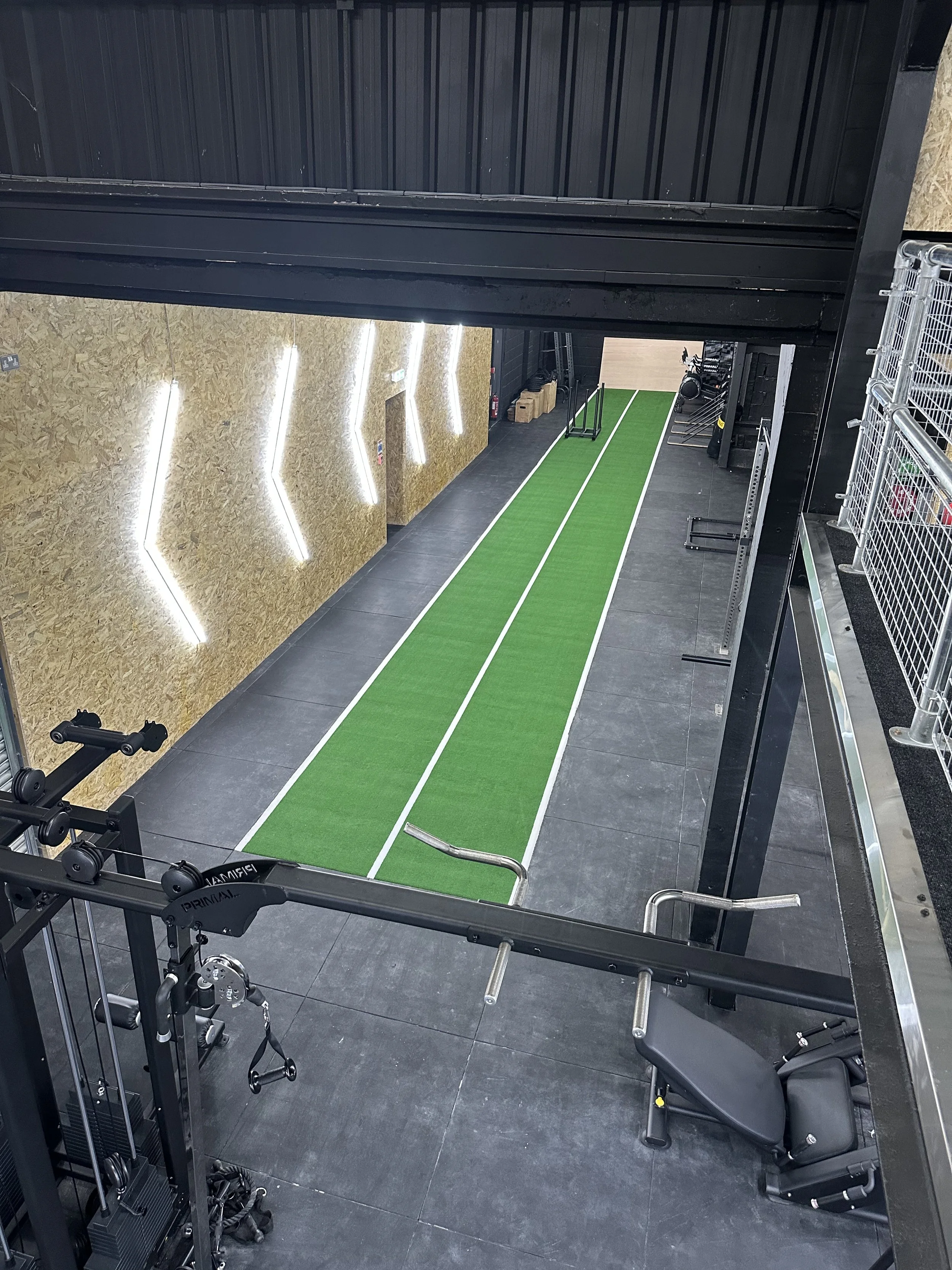 An indoor gym with a green and white running track, black flooring, workout equipment, and wall lighting.