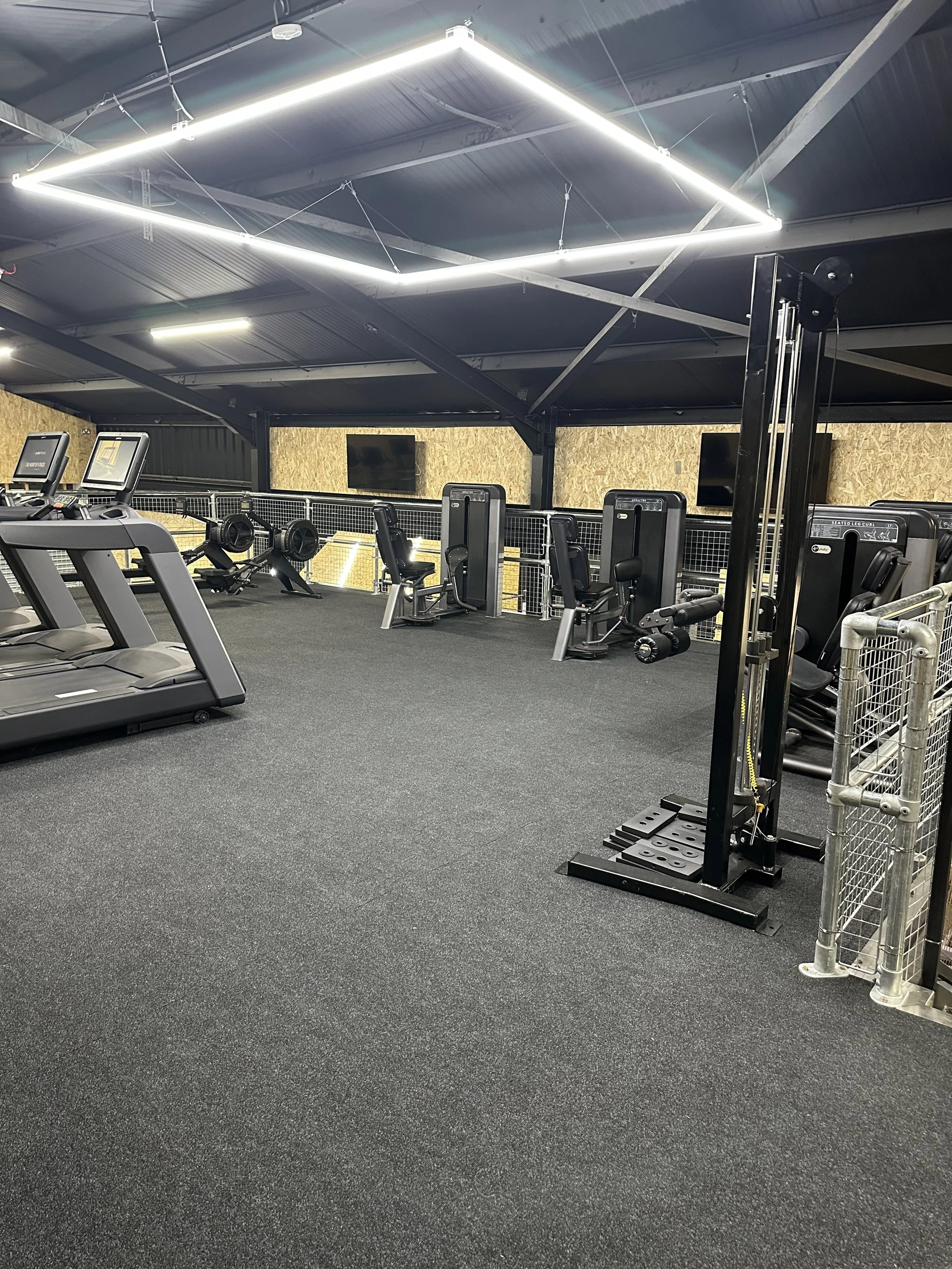 Empty gym with treadmills, weight machines, and TVs on the wall, illuminated by modern rectangular ceiling lights.