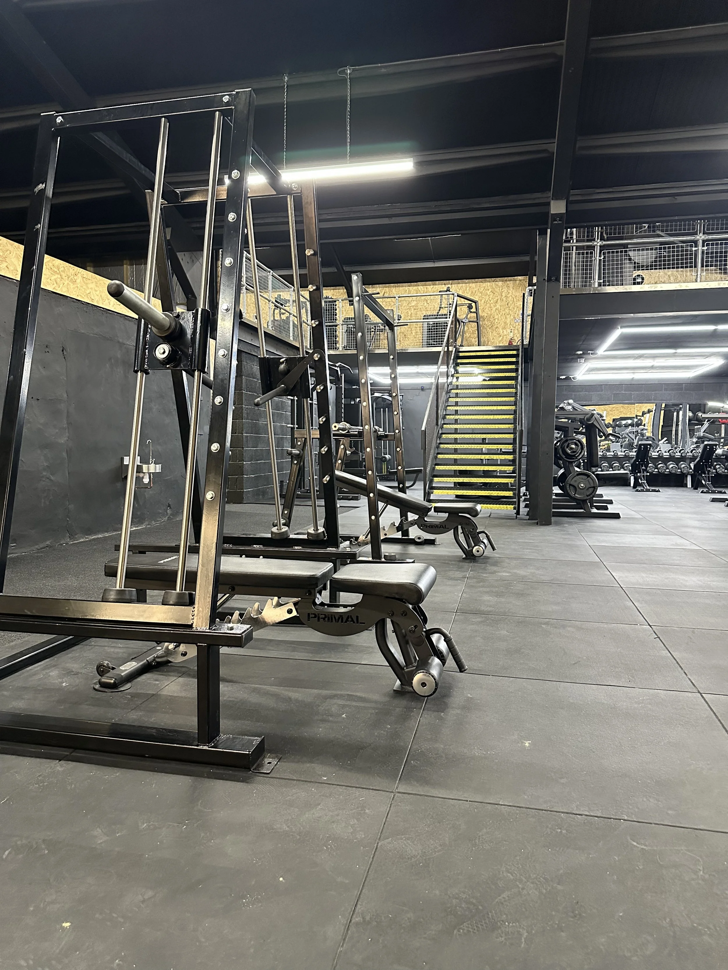 Interior view of a gym with black equipment including a bench and weight racks, a staircase with yellow safety stripes, and a wall with additional weights and machines in the background.