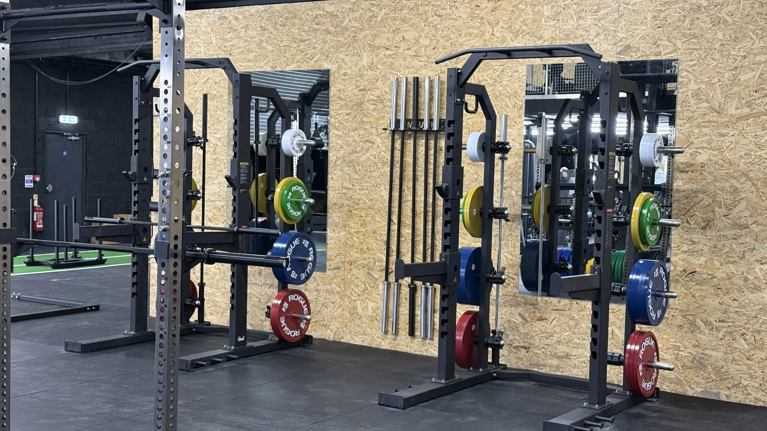 Fitness gym with black weightlifting racks storing colorful weight plates, with a pegboard wall and black rubber flooring.
