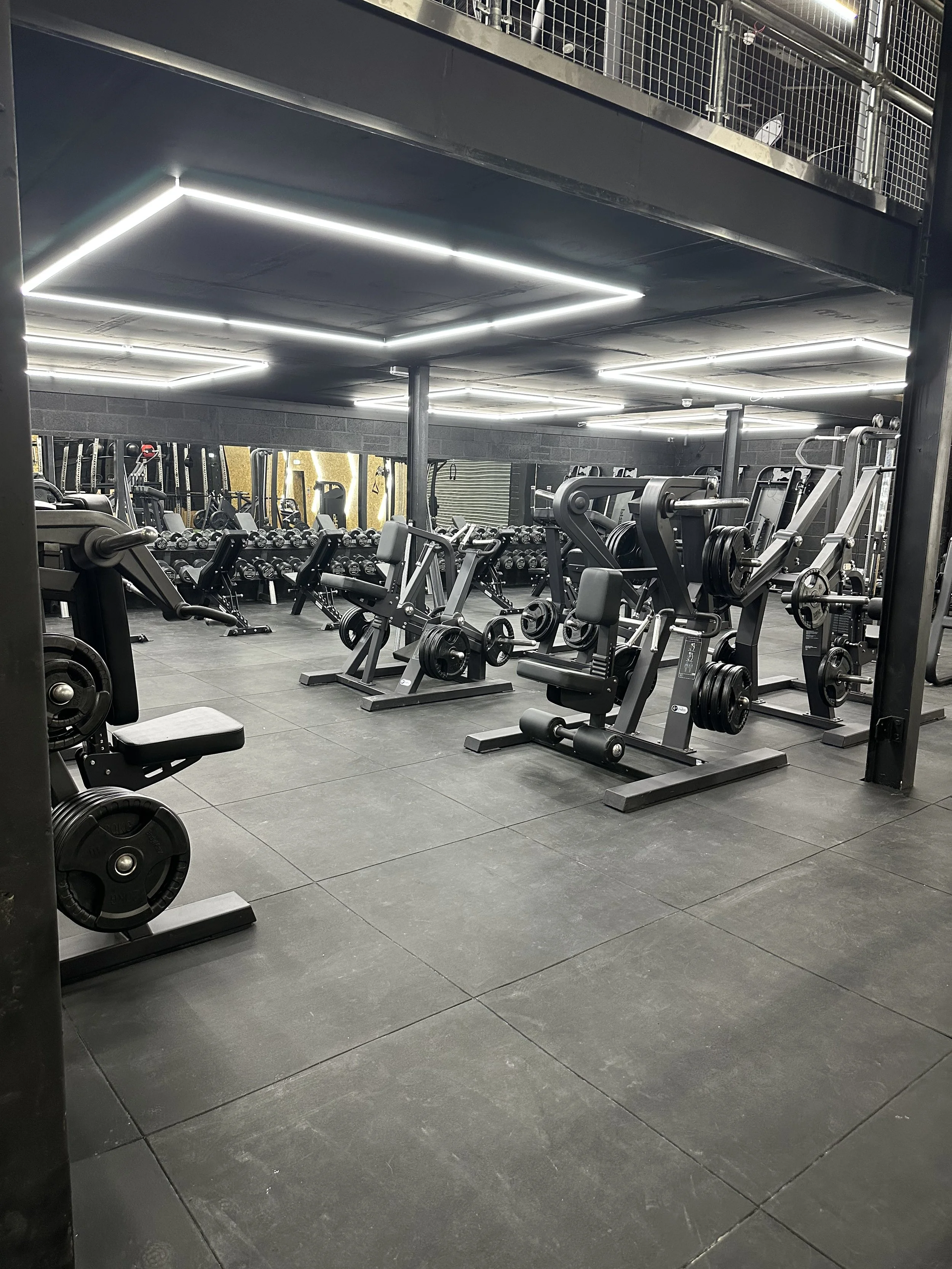 Empty gym with various weightlifting and resistance training equipment, black flooring, and illuminated ceiling panels.