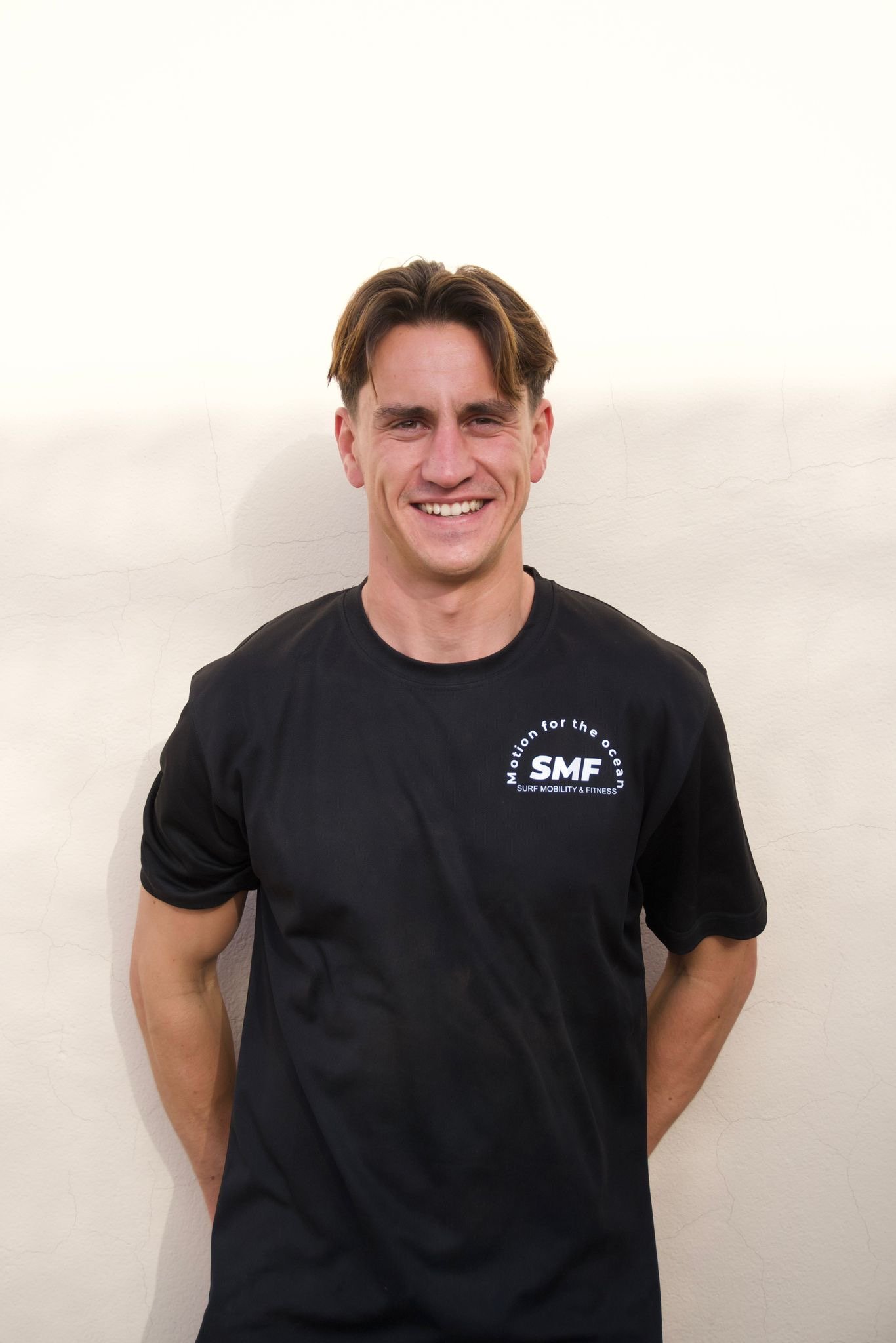A smiling young man with brown hair wearing a black T-shirt with the logo "SMF" and the text "Motion for the ocean" and "Surf Mobility & Fitness" standing against a plain off-white wall.