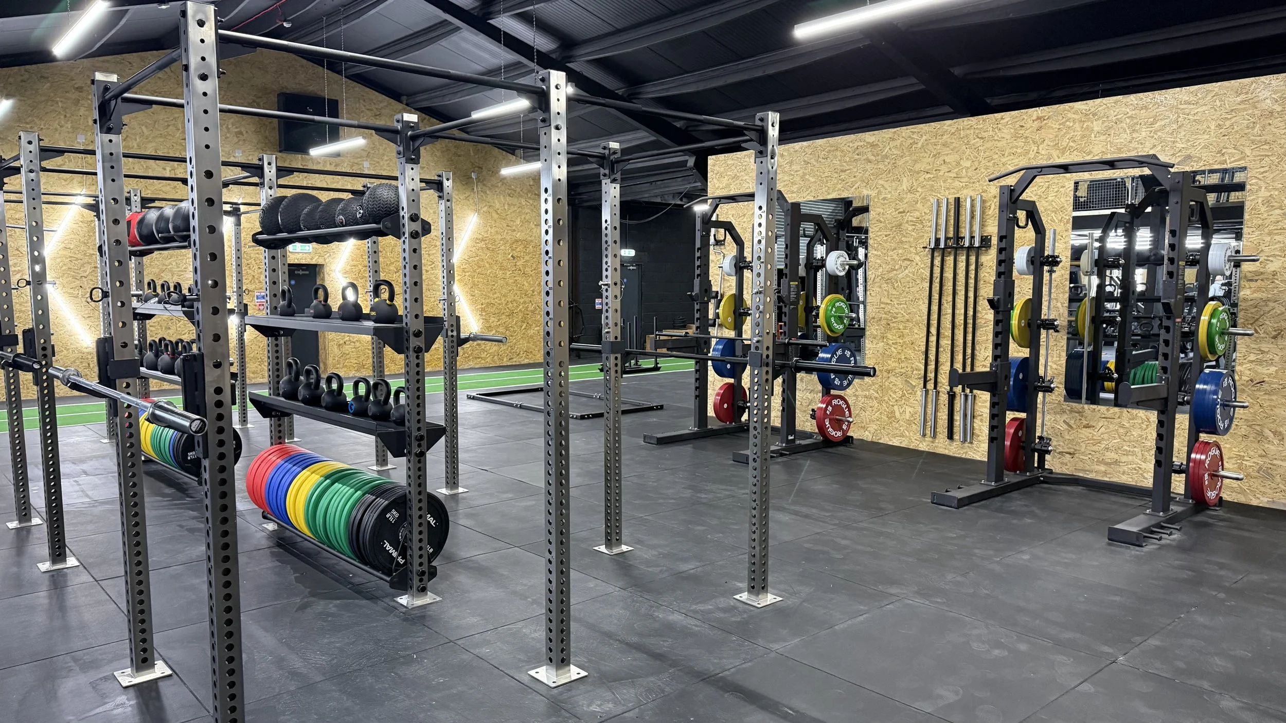 Interior view of a modern gym with various weightlifting equipment, including dumbbells, kettlebells, barbells, and weight plates on racks, set against a yellow textured wall and black rubber flooring.
