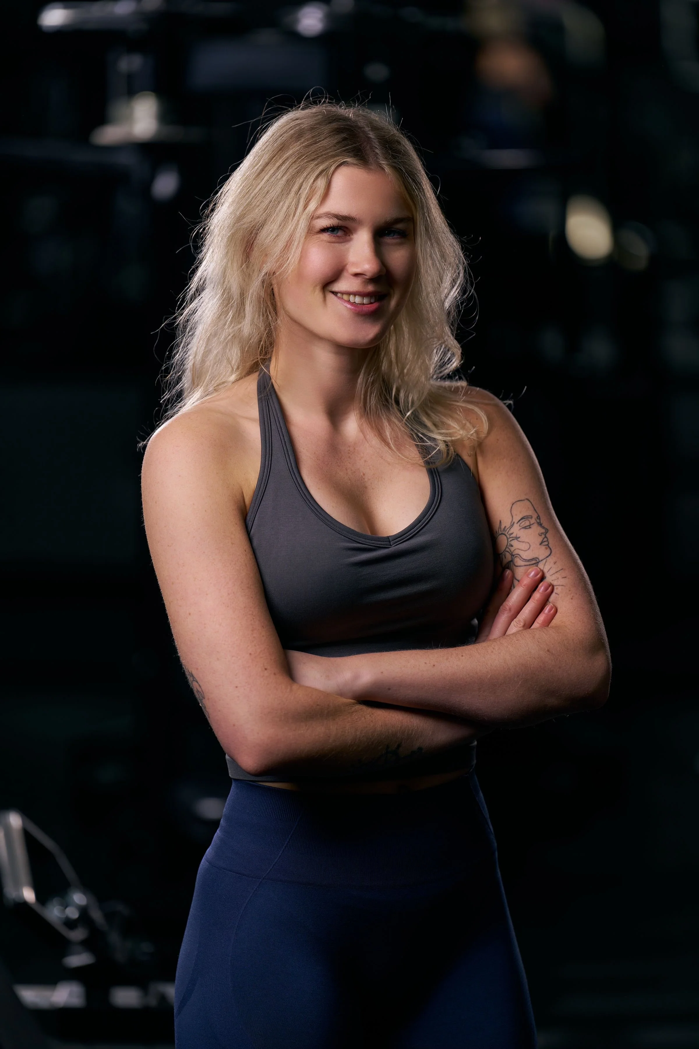 A young woman with blonde hair in workout attire, arms crossed, smiling at the camera in a dimly lit gym.