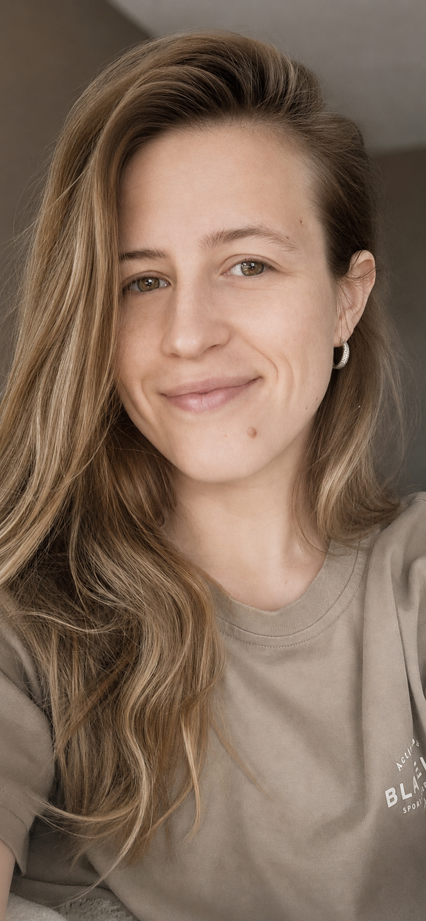 Close-up van een lachende jonge vrouw met lange, golvende lichtbruine haar, lichte huid en oorbellen, draag een beige t-shirt, binnenhuisachtergrond.
