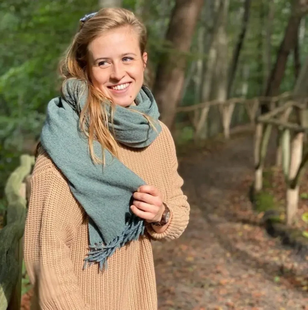 Vrouw in een bos, gekleed in een beige trui en een lichtblauw sjaal, glimlachend en zich vriendelijk verterend.