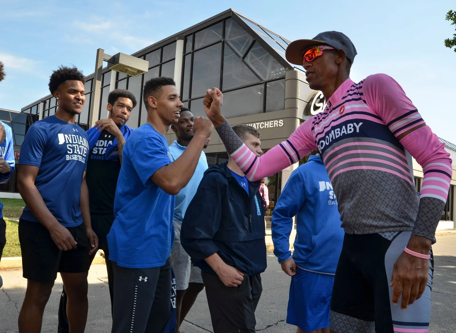 Group of young male athletes in blue Indianapolis State Baseball shirts greet a man in a pink and gray sports shirt with sunglasses outside a building.