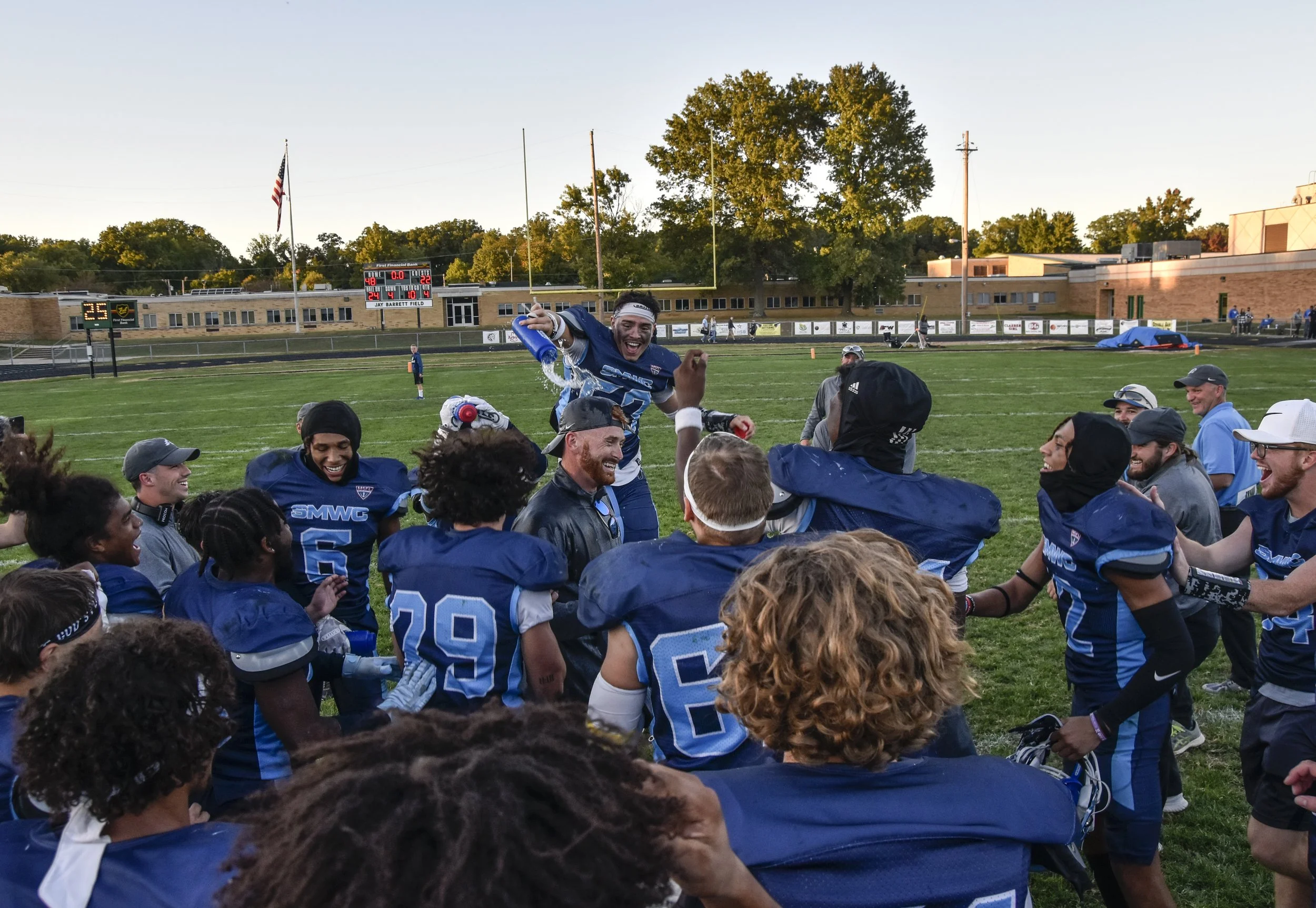 Football team members celebrating on the field after a game, with one player pouring water over a teammate's head, during sunset at the high school stadium.