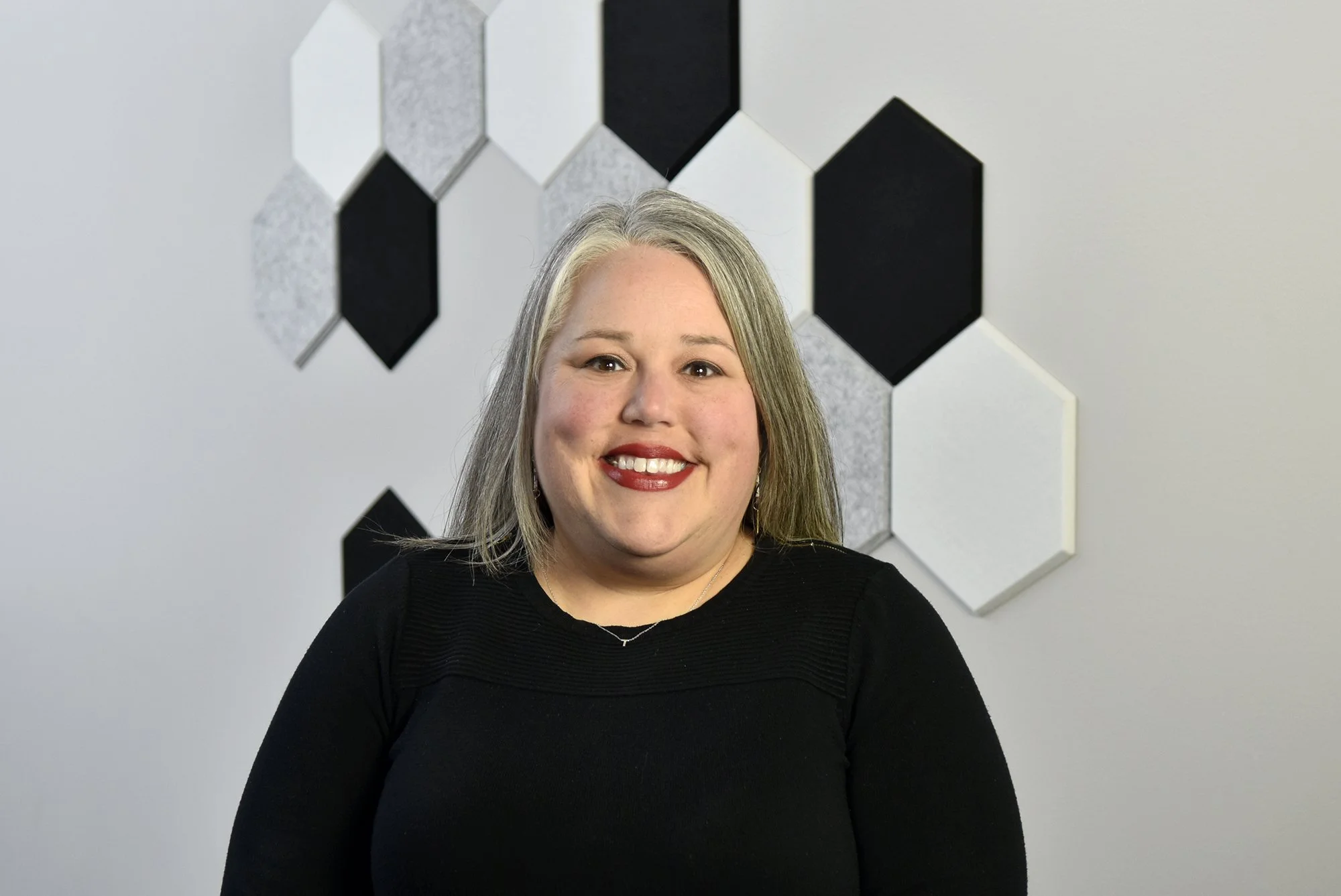 A smiling woman with shoulder-length blonde hair, wearing a black top, standing in front of a wall with geometric black, white, and gray hexagon-shaped wall decorations.