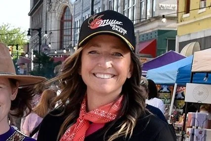 Smiling woman at outdoor market, wearing a Miller Lite cap and red bandana, with colorful tents and buildings in background.